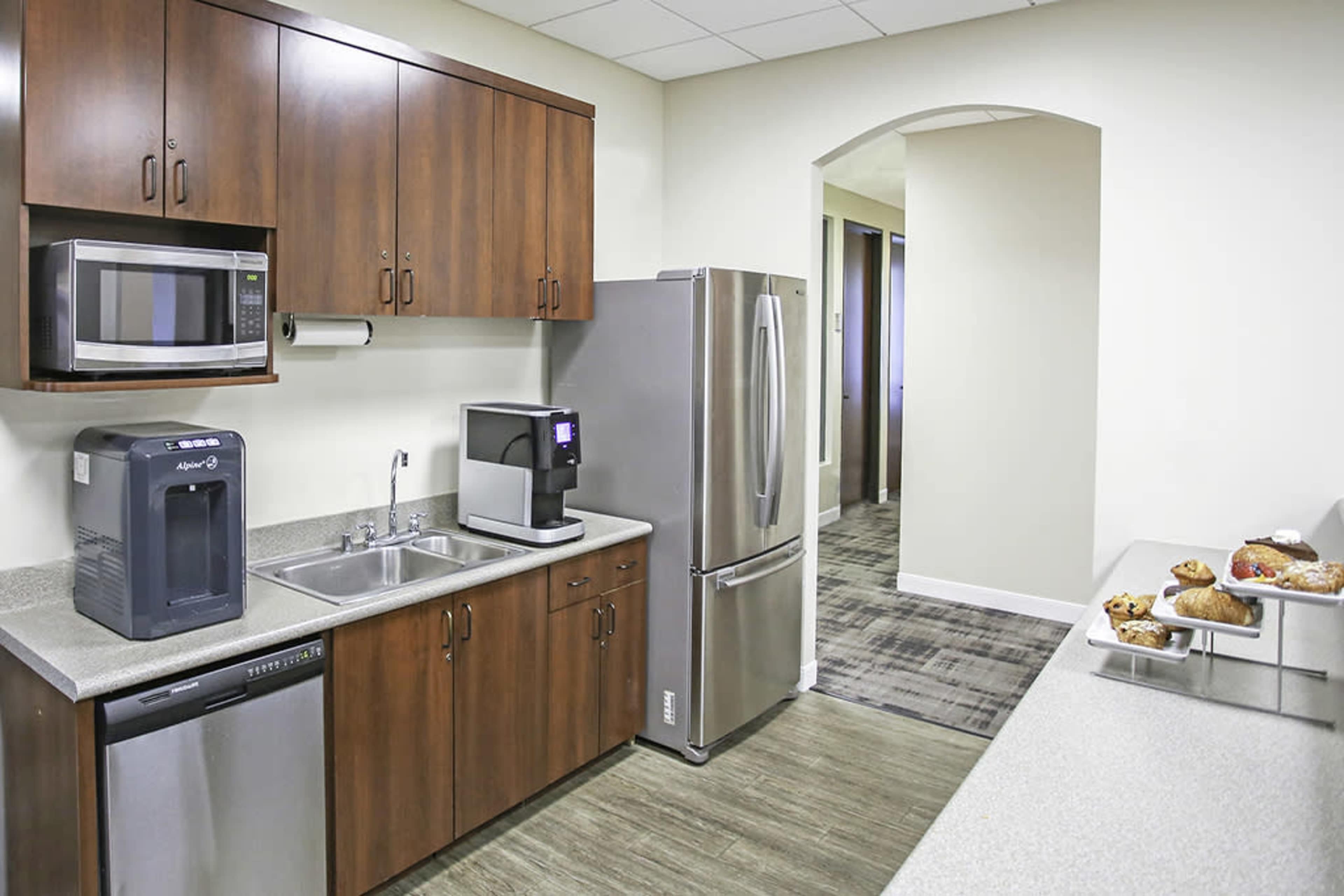 A modern kitchen area equipped with wooden cabinets, a stainless steel refrigerator, a microwave, a coffee machine, and pastries on a counter.