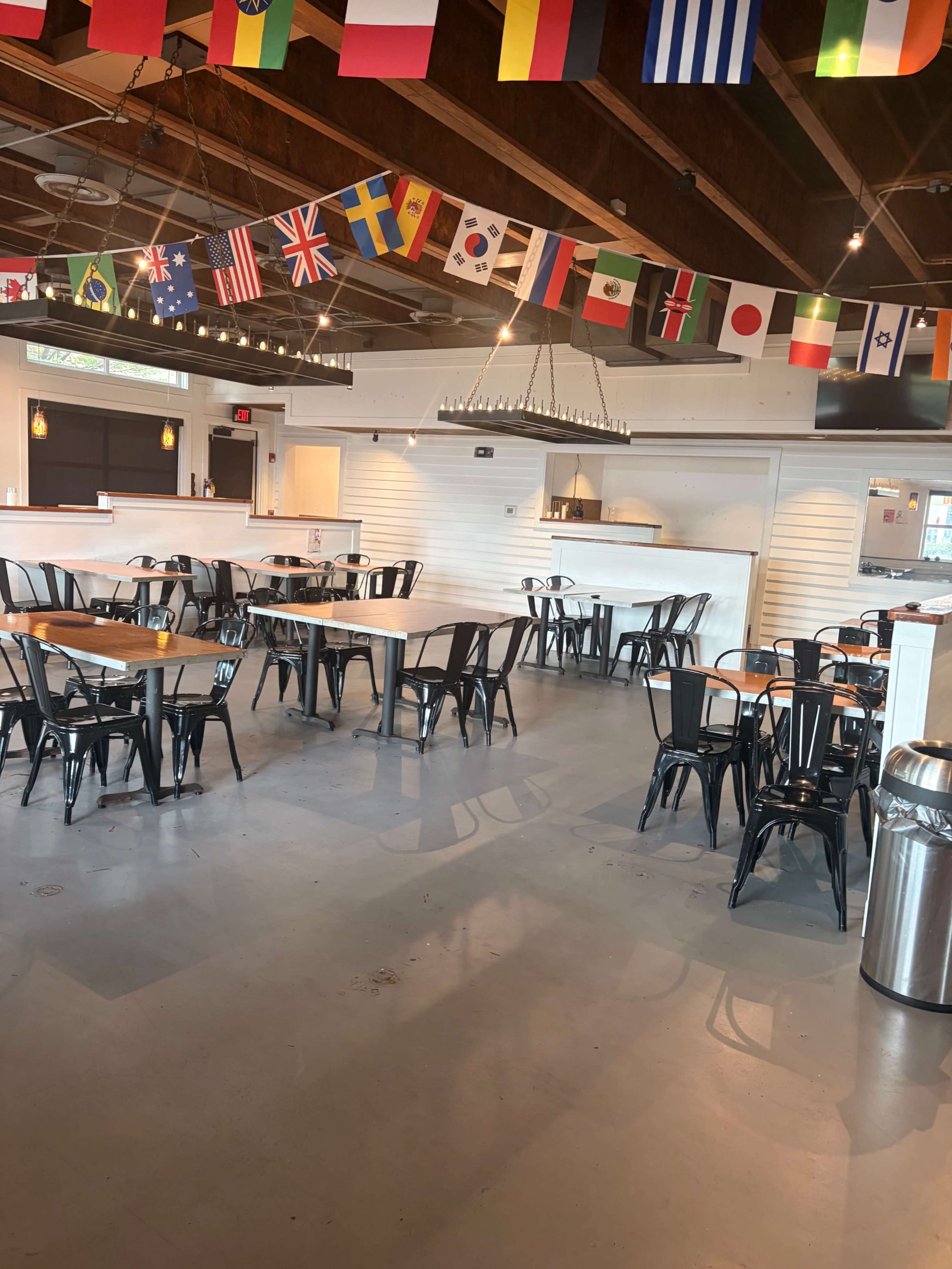 The image shows a spacious dining area with black metal chairs and tables, decorated with various international flags hanging from the ceiling.