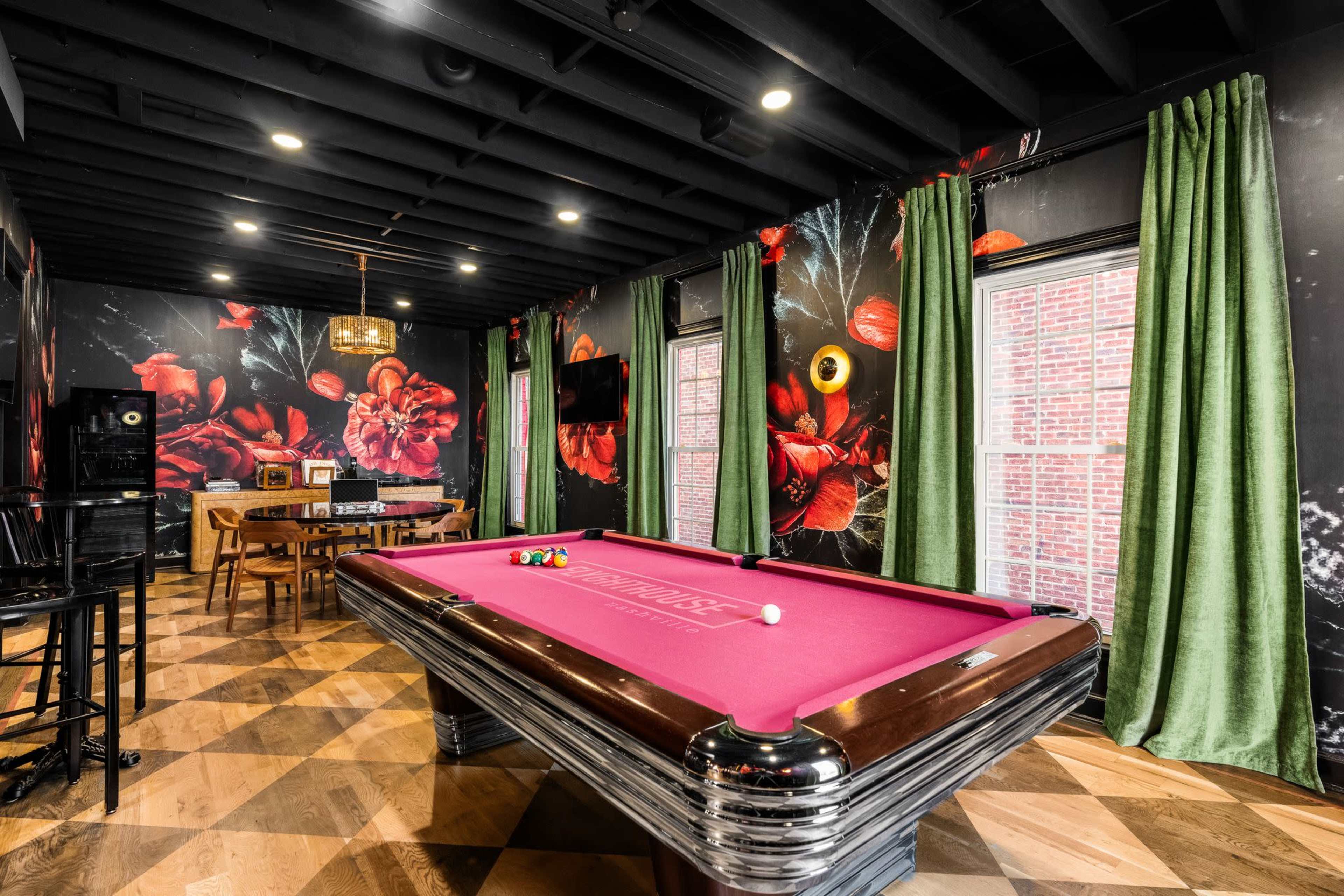 A game room featuring a pink pool table, dark floral wallpaper, and green curtains, with wooden flooring and a dining table in the background.