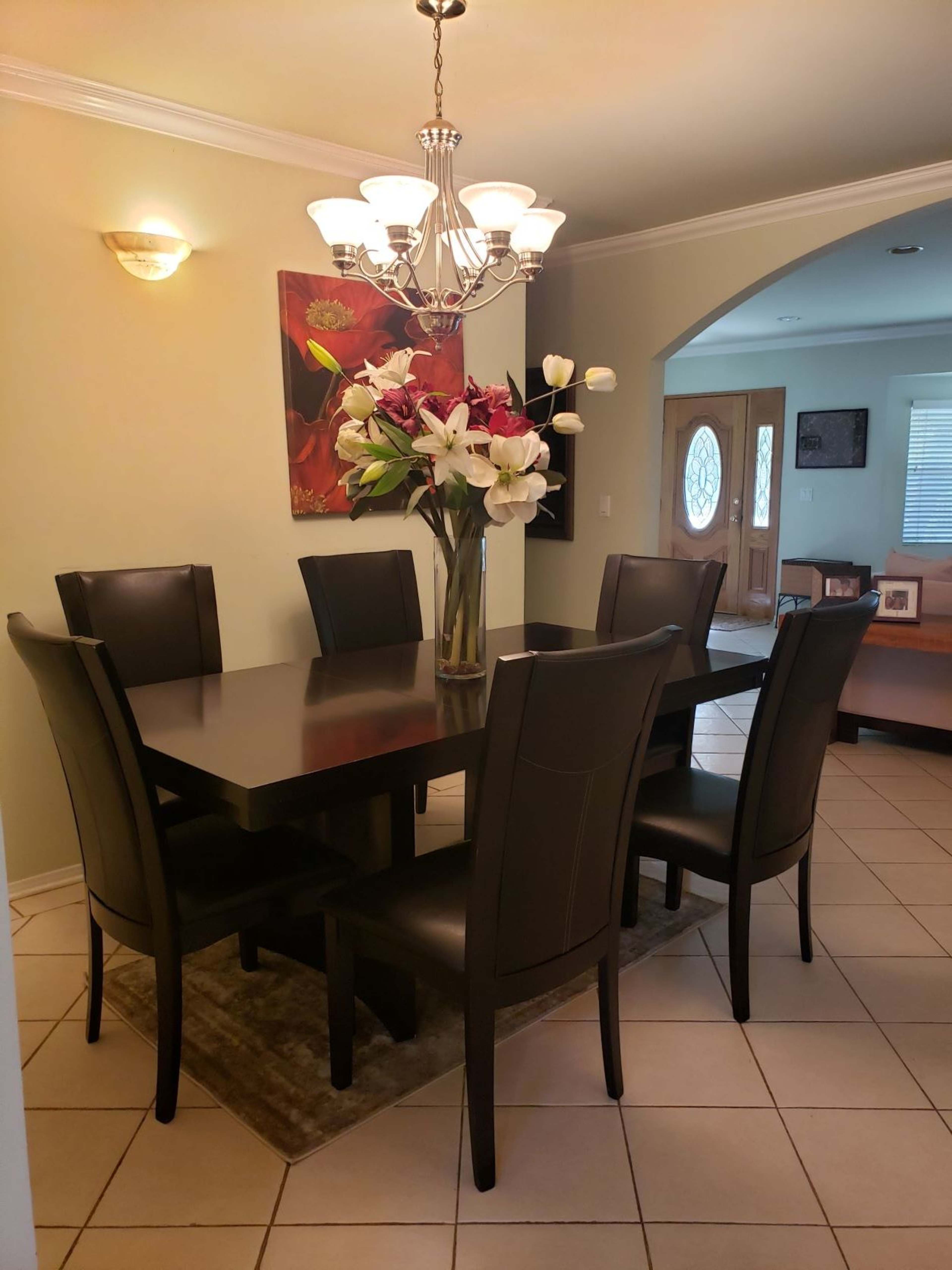 A modern dining room features a rectangular table surrounded by six leather chairs, with a large floral arrangement in the center and a chandelier above.