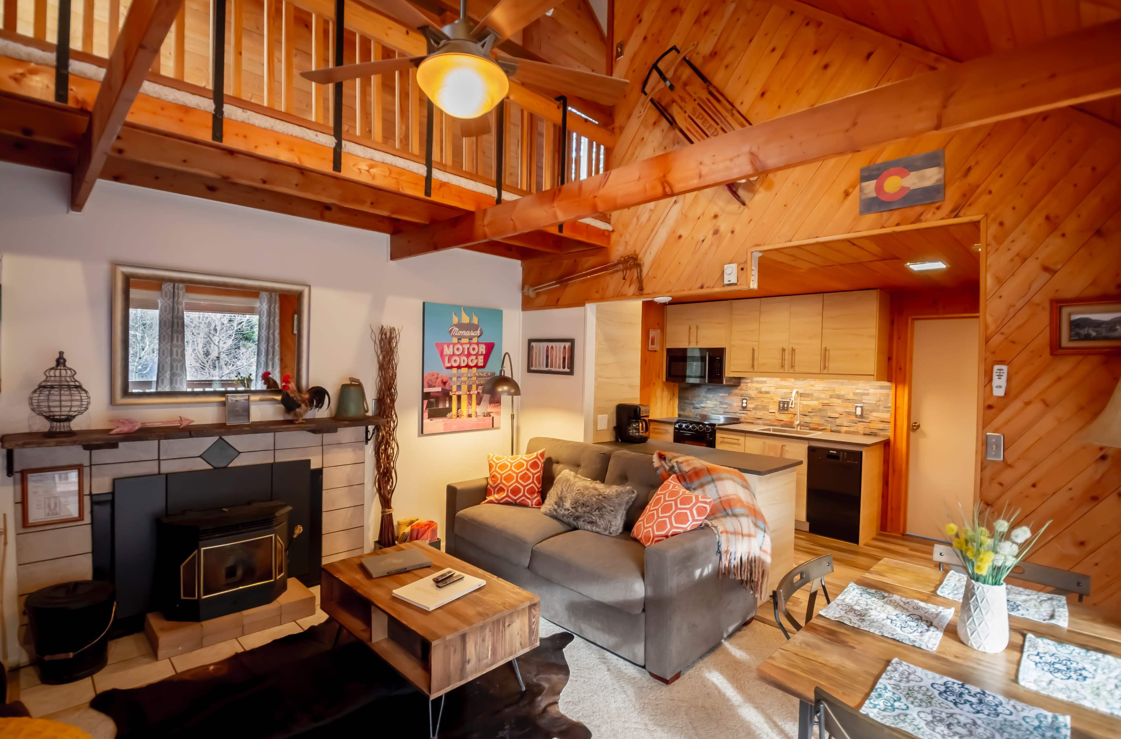 A cozy cabin interior features a wooden ceiling, a fireplace, and an open kitchen with modern appliances.