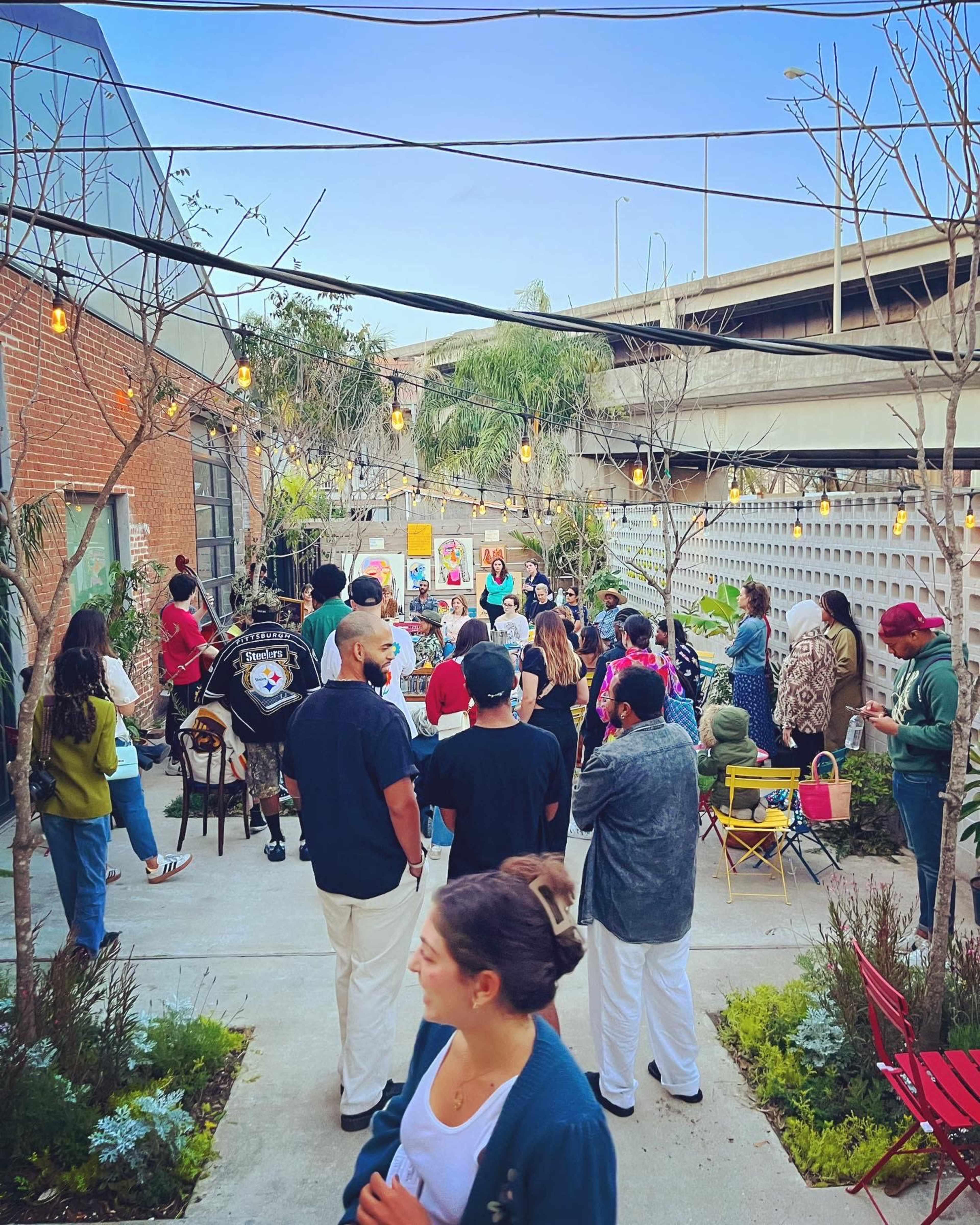 A crowd gathers in an outdoor space with string lights and greenery, while a speaker addresses the group.