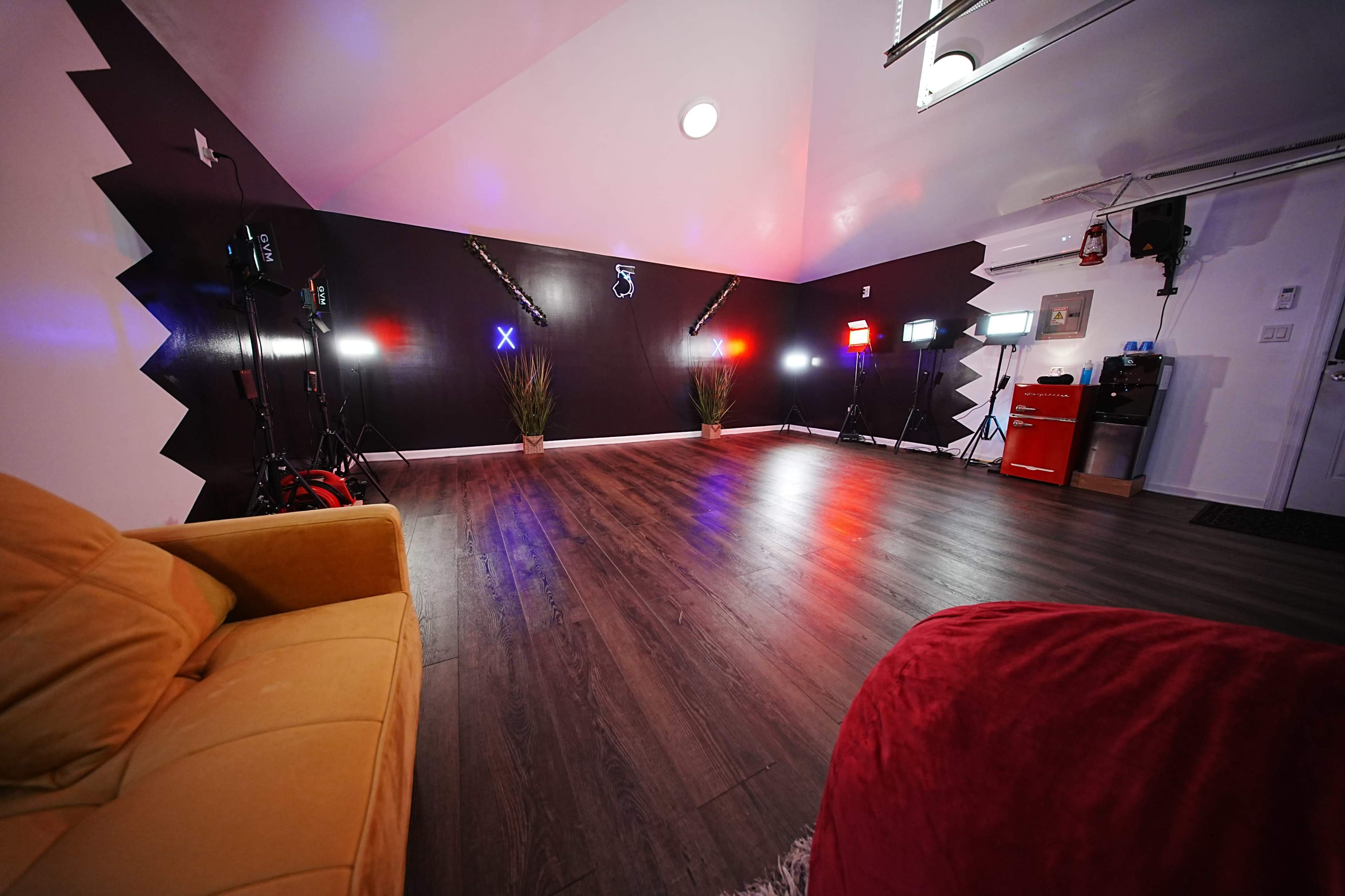 The image shows a spacious room featuring a wooden floor, a yellow sofa, colorful lighting equipment, and potted plants against a dark wall.