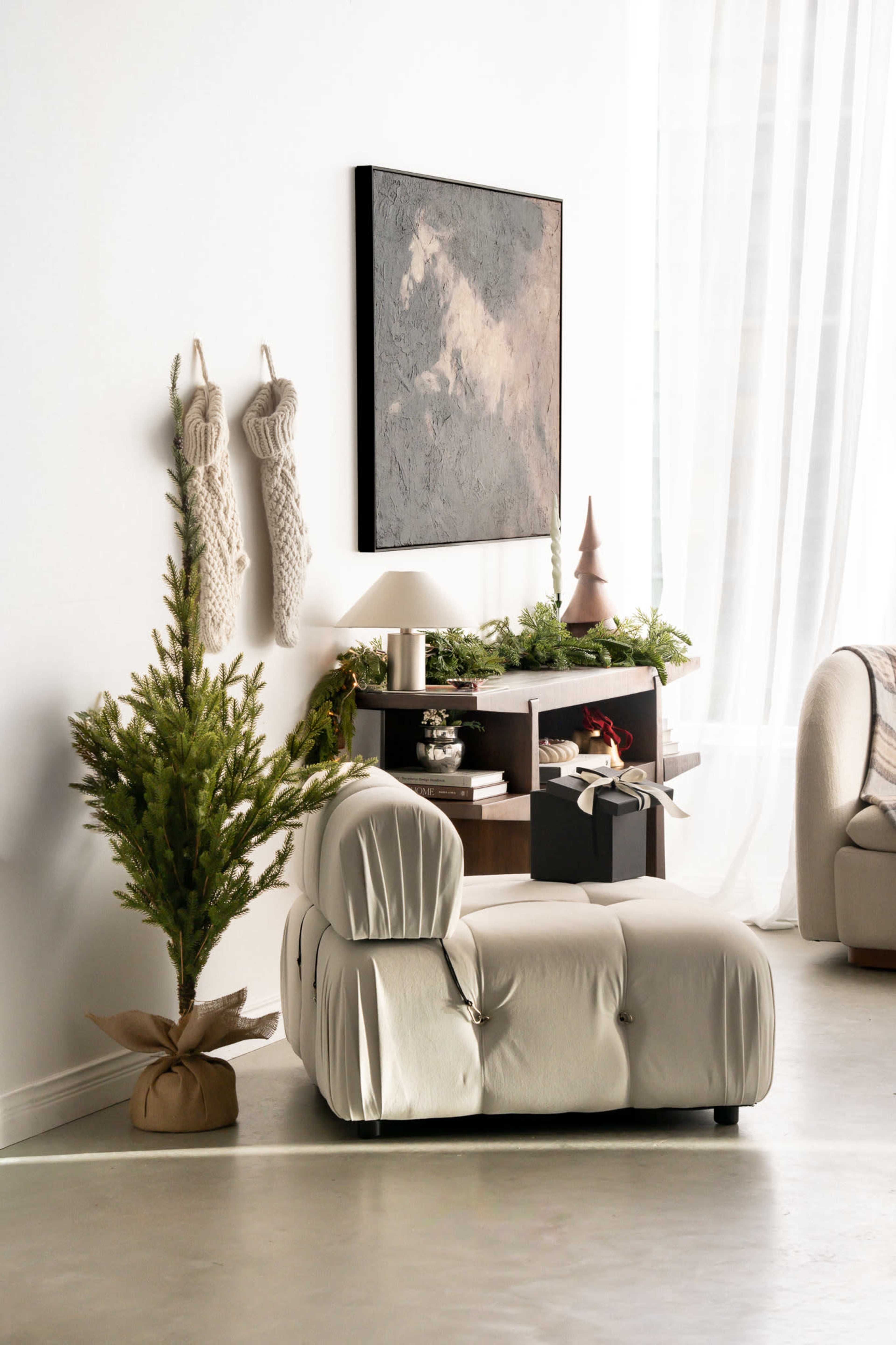 The image shows a modern living room with a cream-colored chaise lounge, a small decorated Christmas tree, and a shelf adorned with holiday decor and gifts against a white wall.