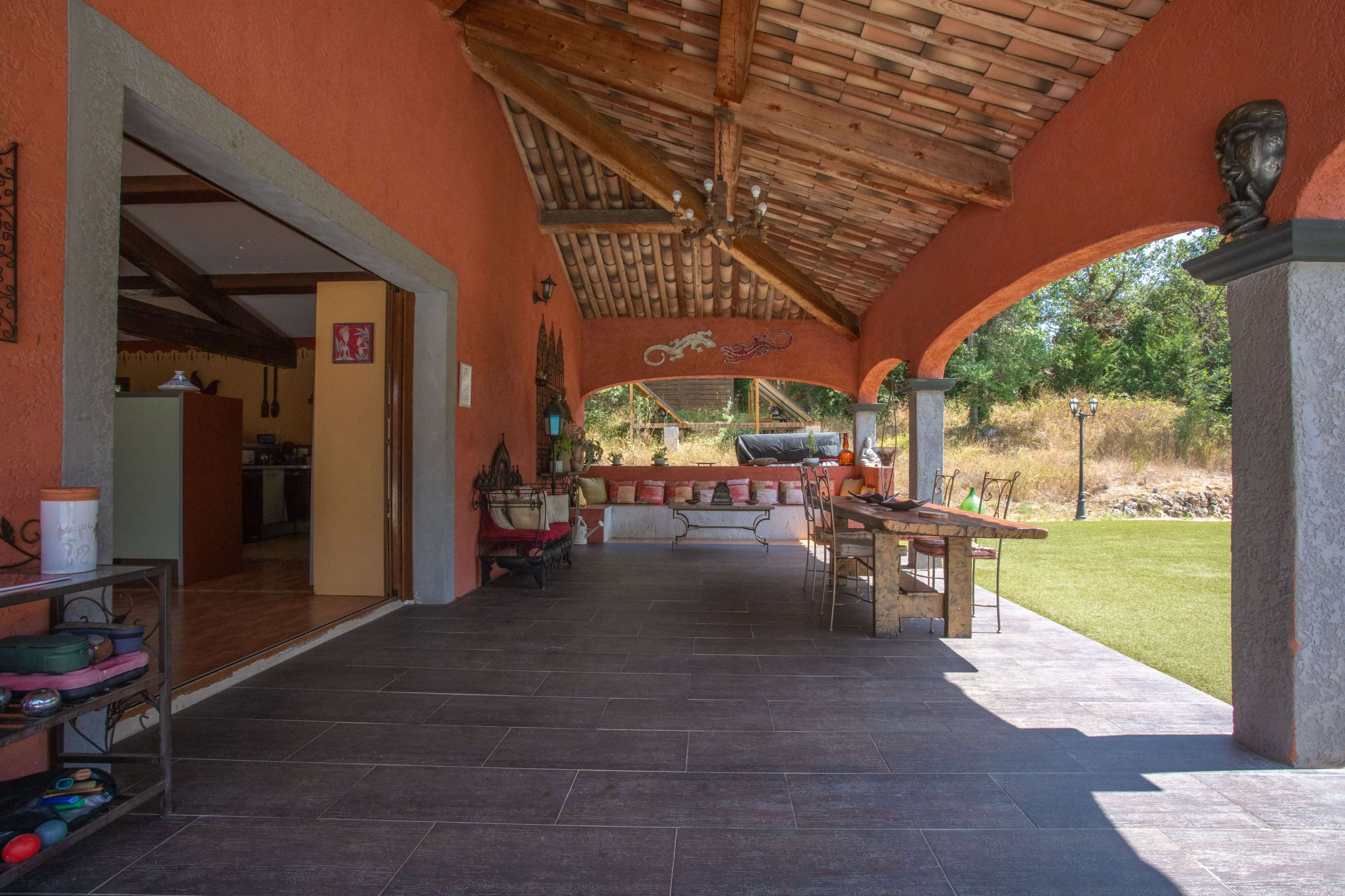 The image shows a spacious outdoor patio with a sloped wooden roof, featuring tables and chairs, and surrounded by greenery.