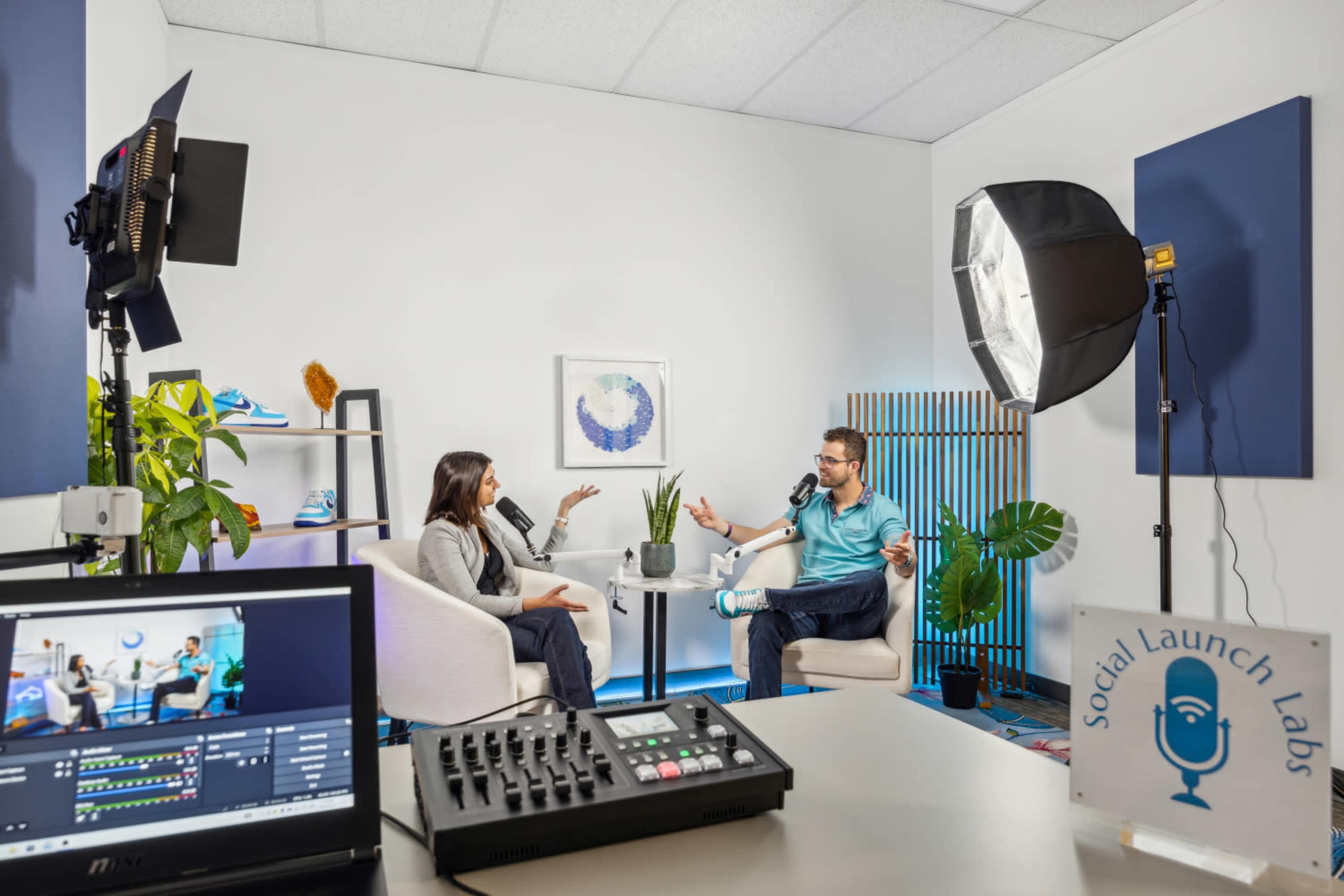 Two individuals are seated in a modern studio setting, engaging in a conversation while recording on a video production setup that includes cameras and lighting equipment.