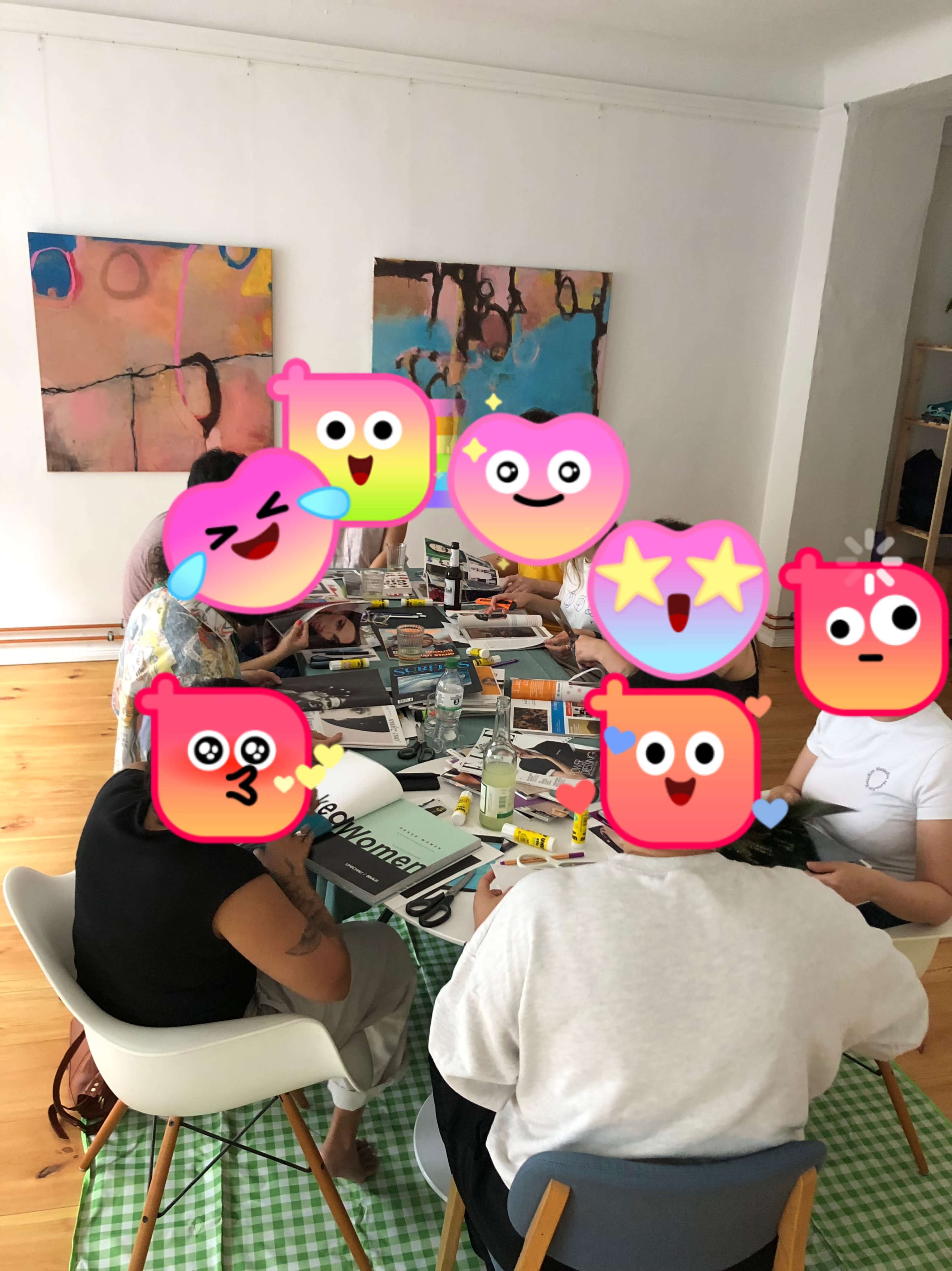 A group of people is seated around a table covered with various art supplies and drinks in a brightly lit room with colorful abstract art on the walls.