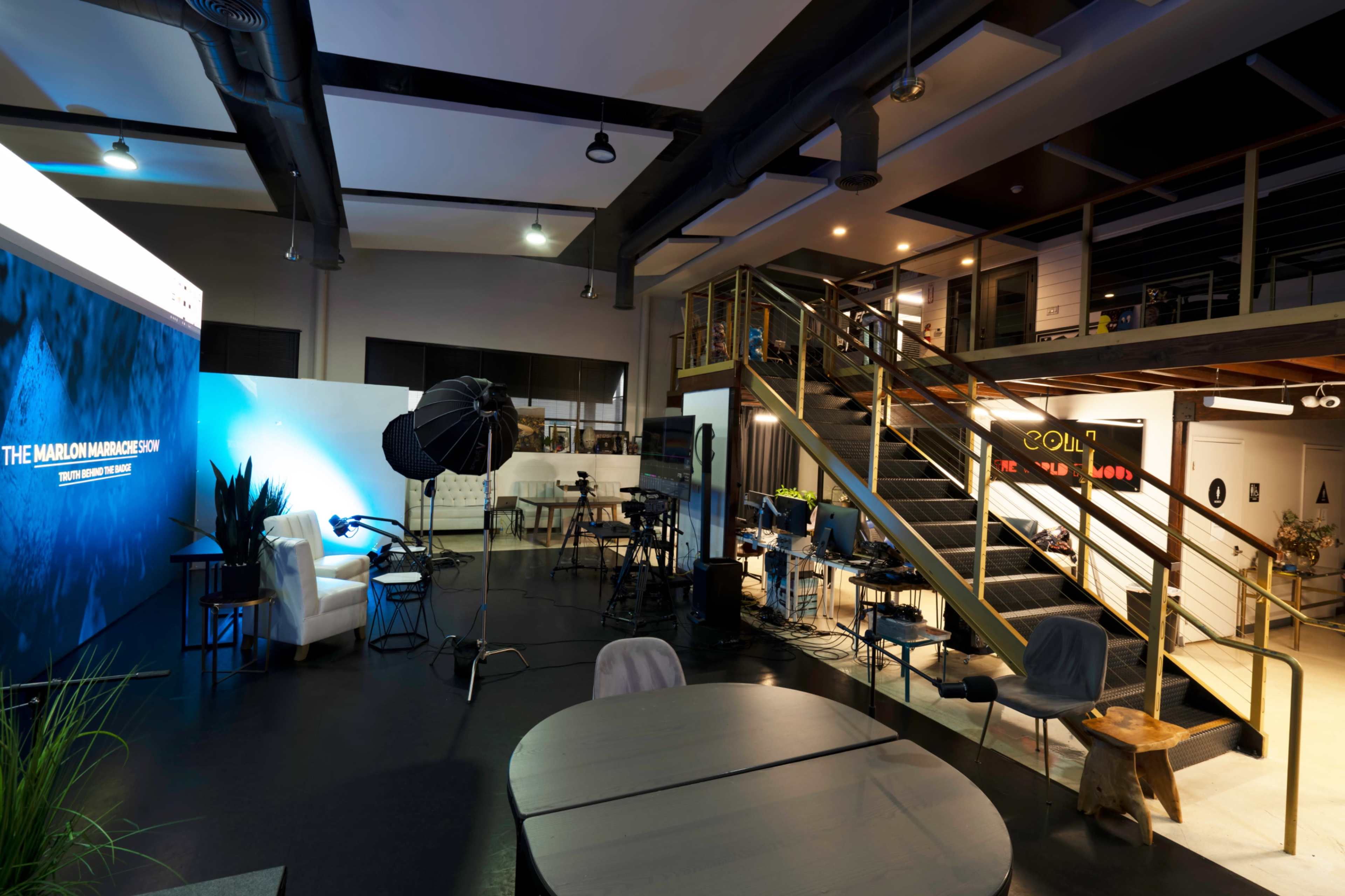 The image shows a modern interior space featuring a staircase, seating areas, and equipment for media production.