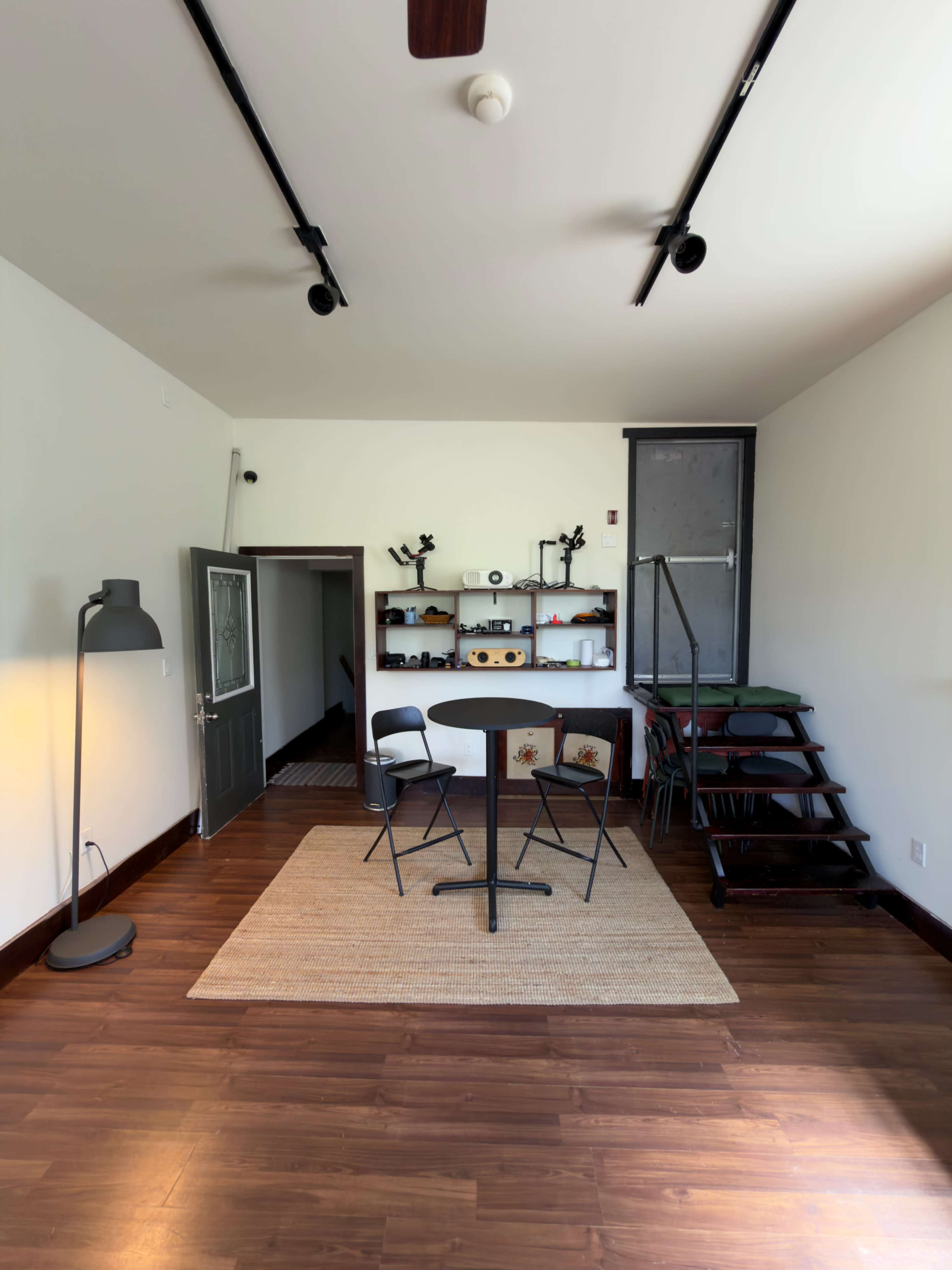 The image shows a modern, minimalistic room featuring a small table with three chairs, a standing lamp, and a staircase leading to a loft area, all against a backdrop of wooden flooring and white walls.
