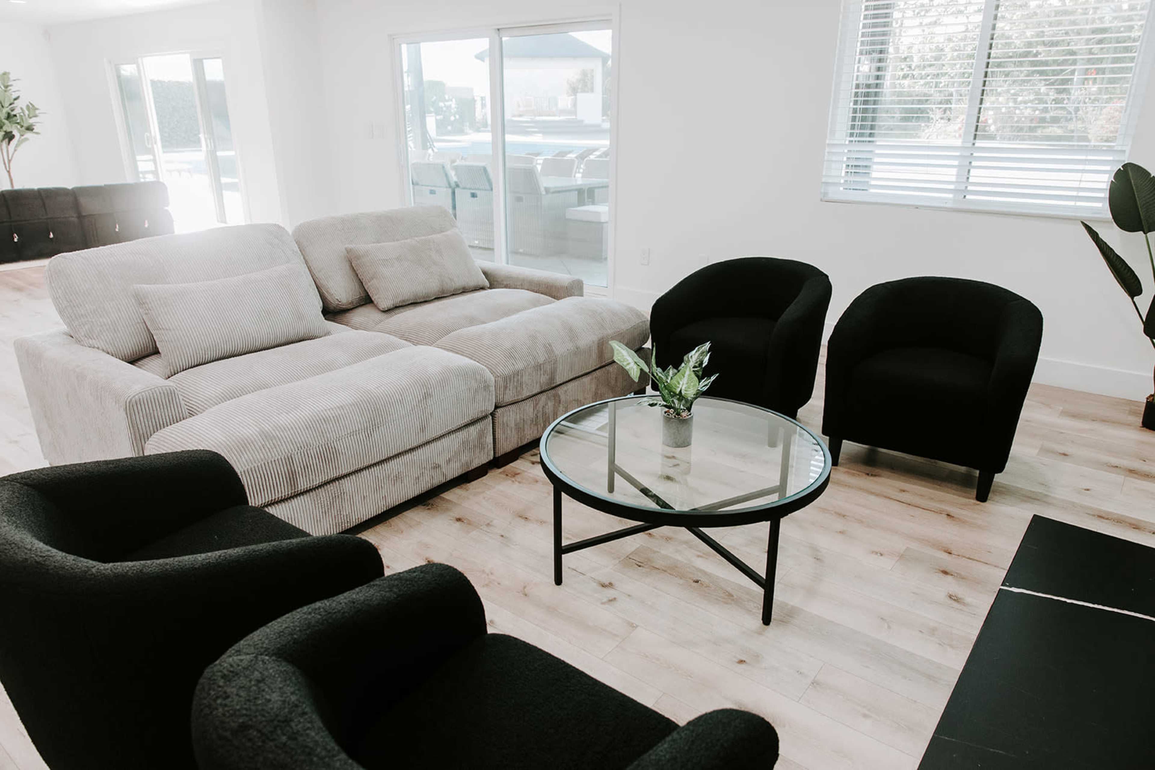 A modern living room features a sectional sofa, two black chairs, a round glass coffee table, and large windows allowing natural light.