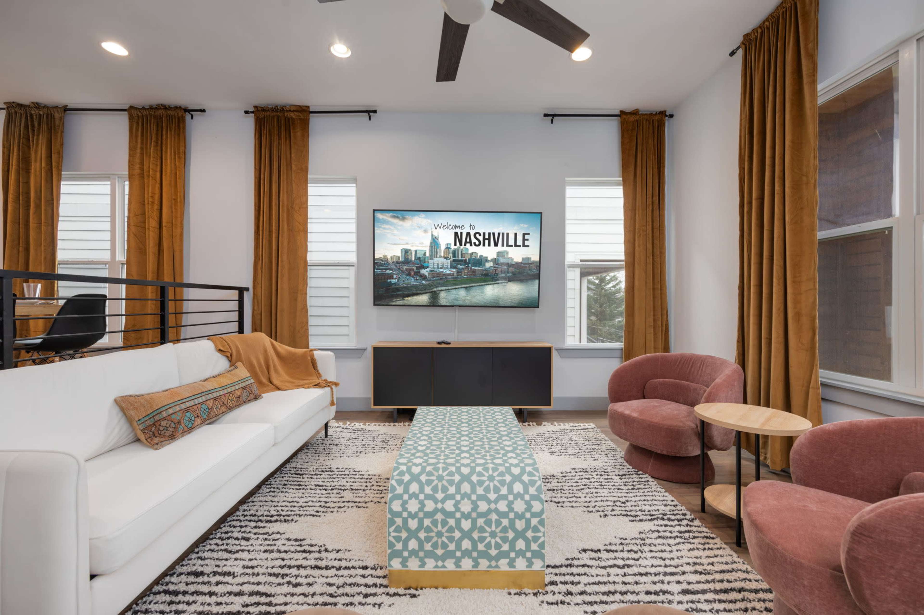 A modern living room with a white sofa, a patterned blue coffee table, two pink chairs, and a large wall-mounted TV displaying "Welcome to Nashville."