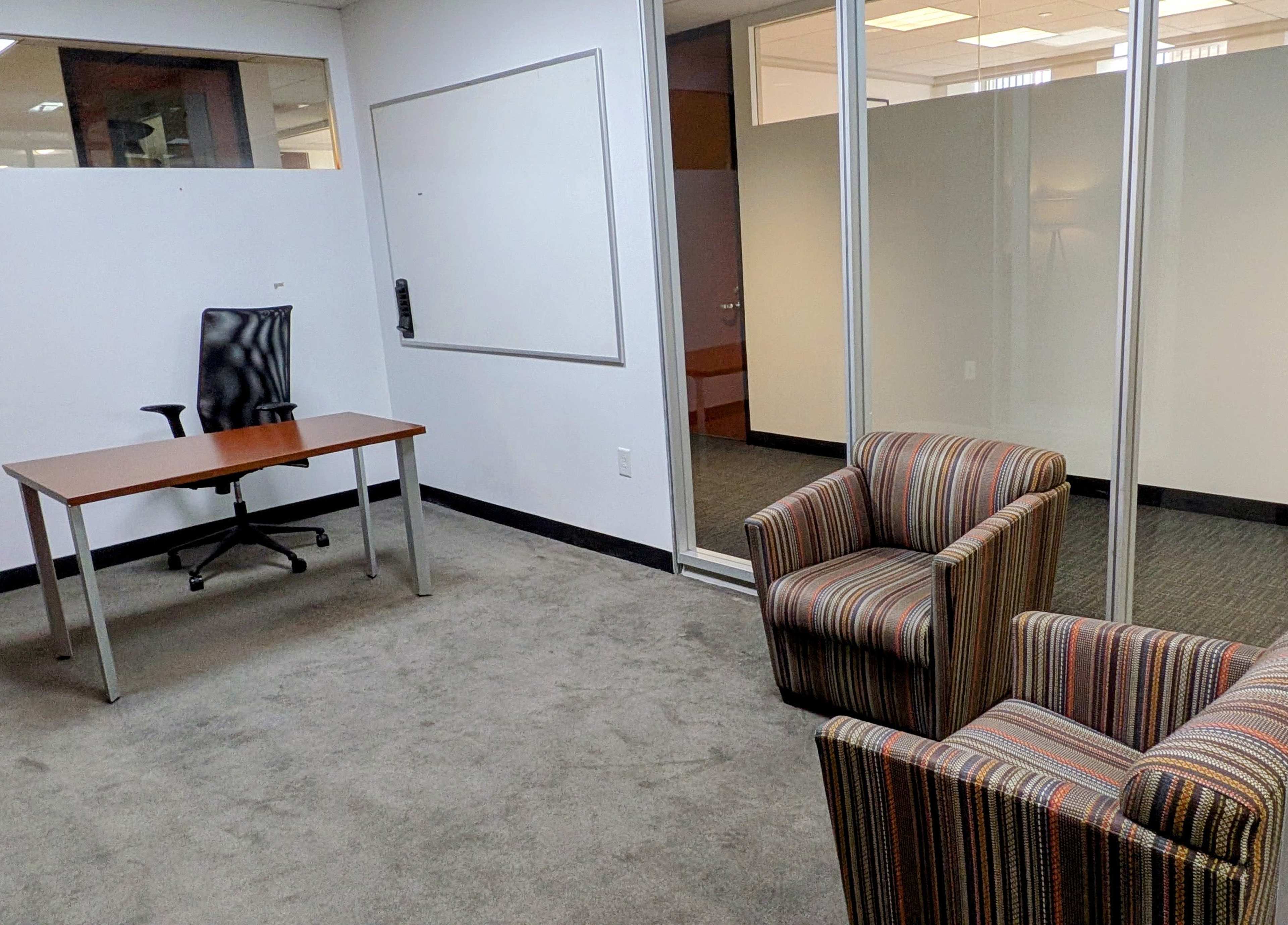 The image shows a small office space featuring a desk and a chair, along with two striped armchairs in a carpeted area.