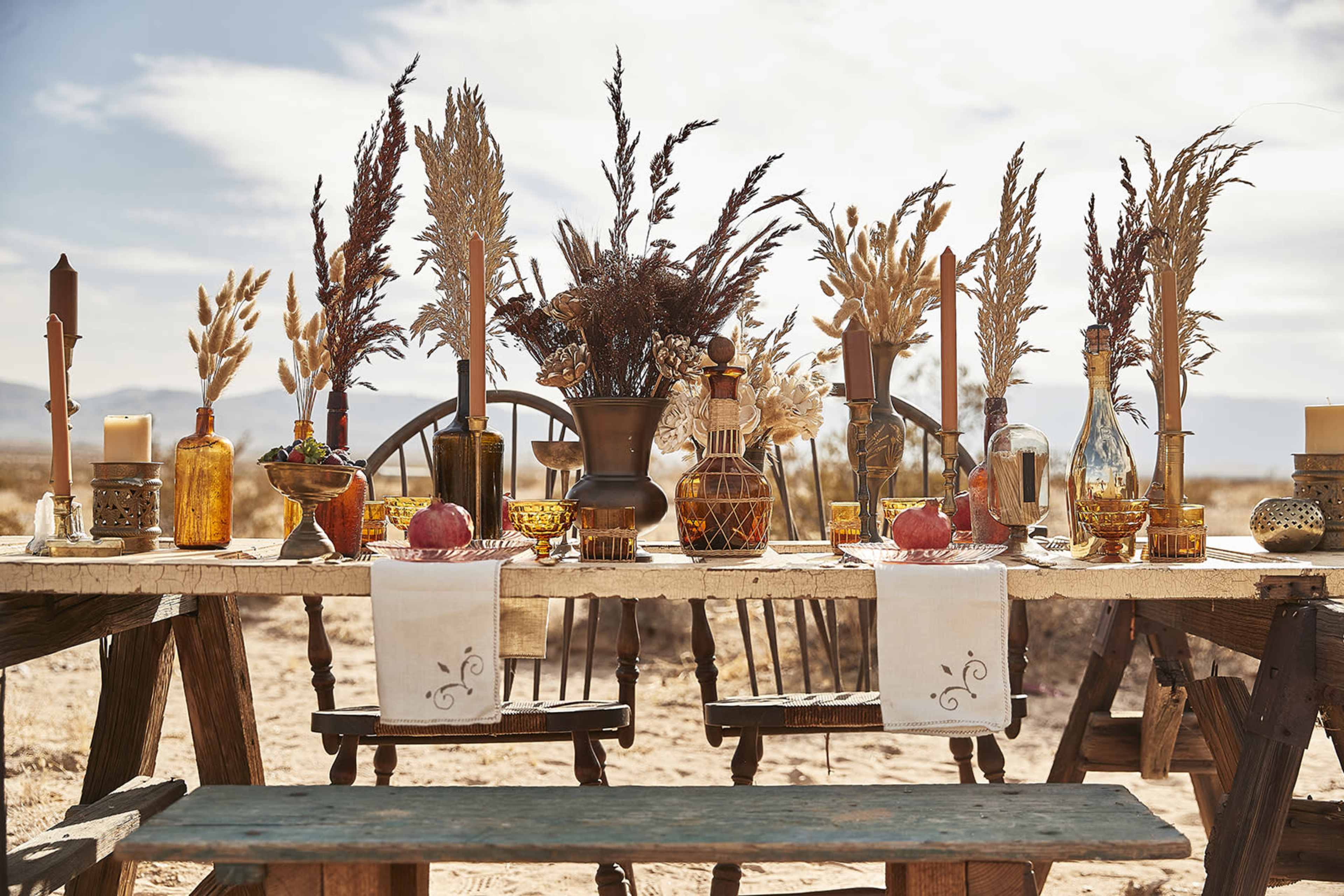 A long wooden table is set outdoors with various decorative items, including glass bottles, candles, and dried floral arrangements in amber tones.