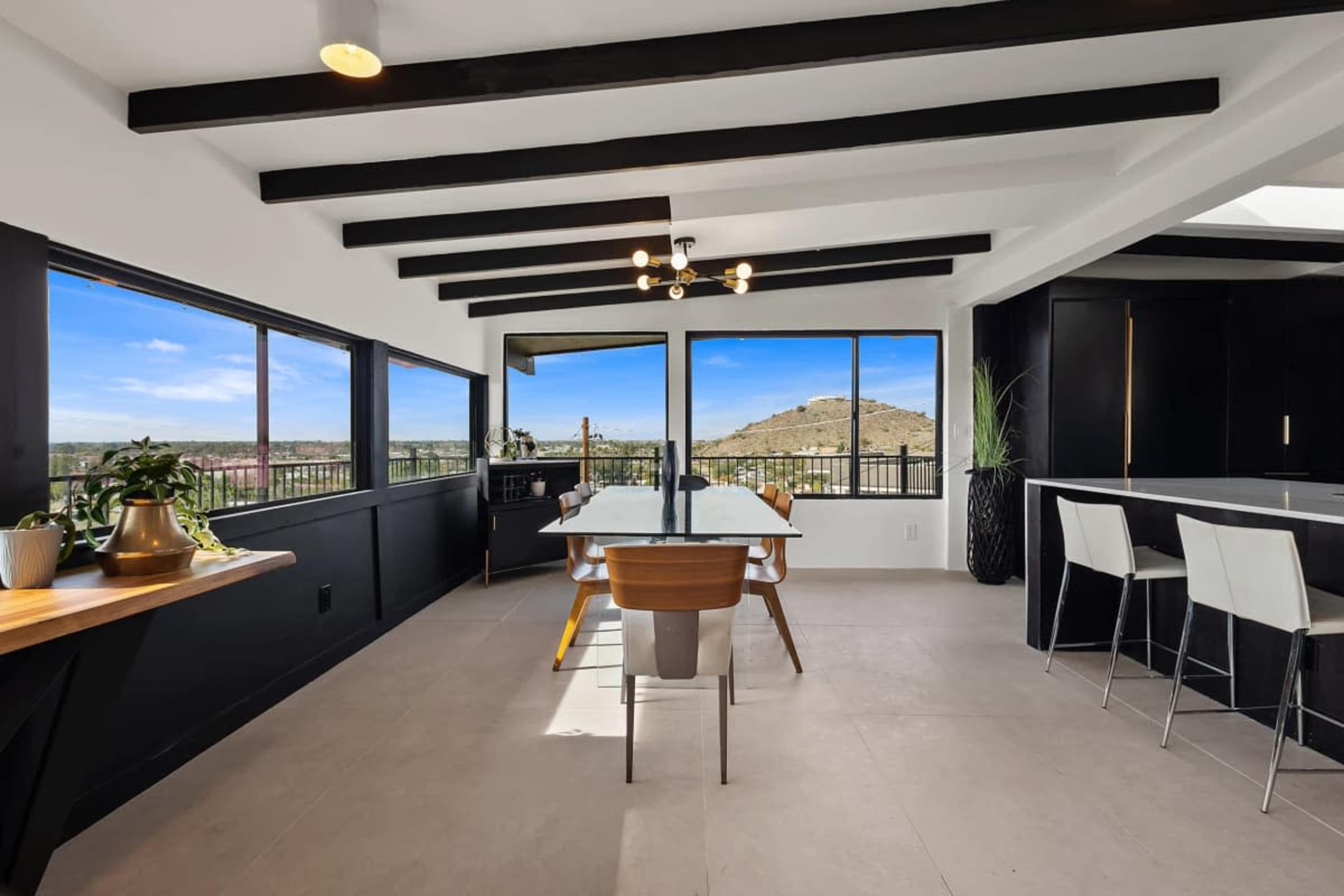 A modern dining area features a long table with wooden chairs, large windows offering views of a hilly landscape, and a sleek kitchen bar.