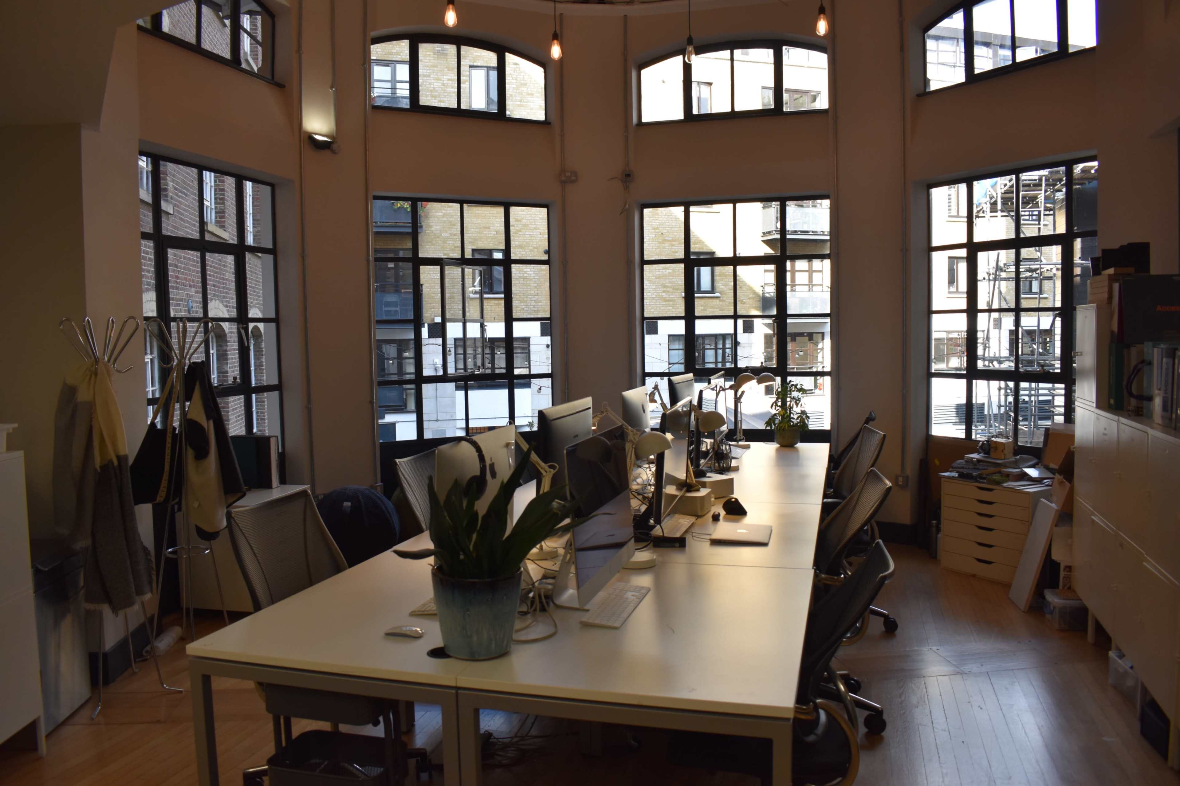 The image shows a modern office space with multiple desks equipped with computers and large windows that provide natural light.