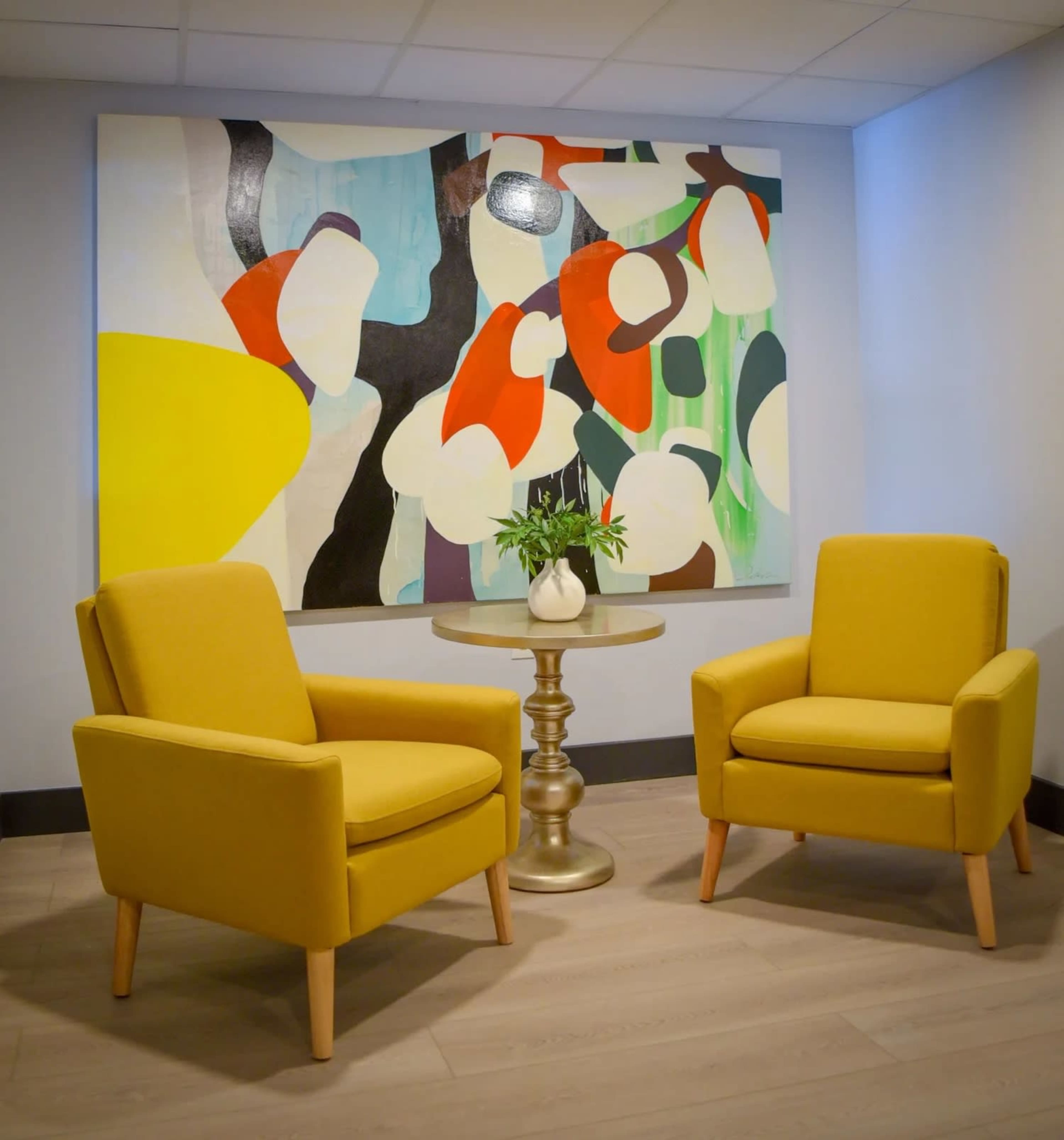Two yellow armchairs are positioned facing each other in a room, with a round gold table in between them, under a large abstract painting.