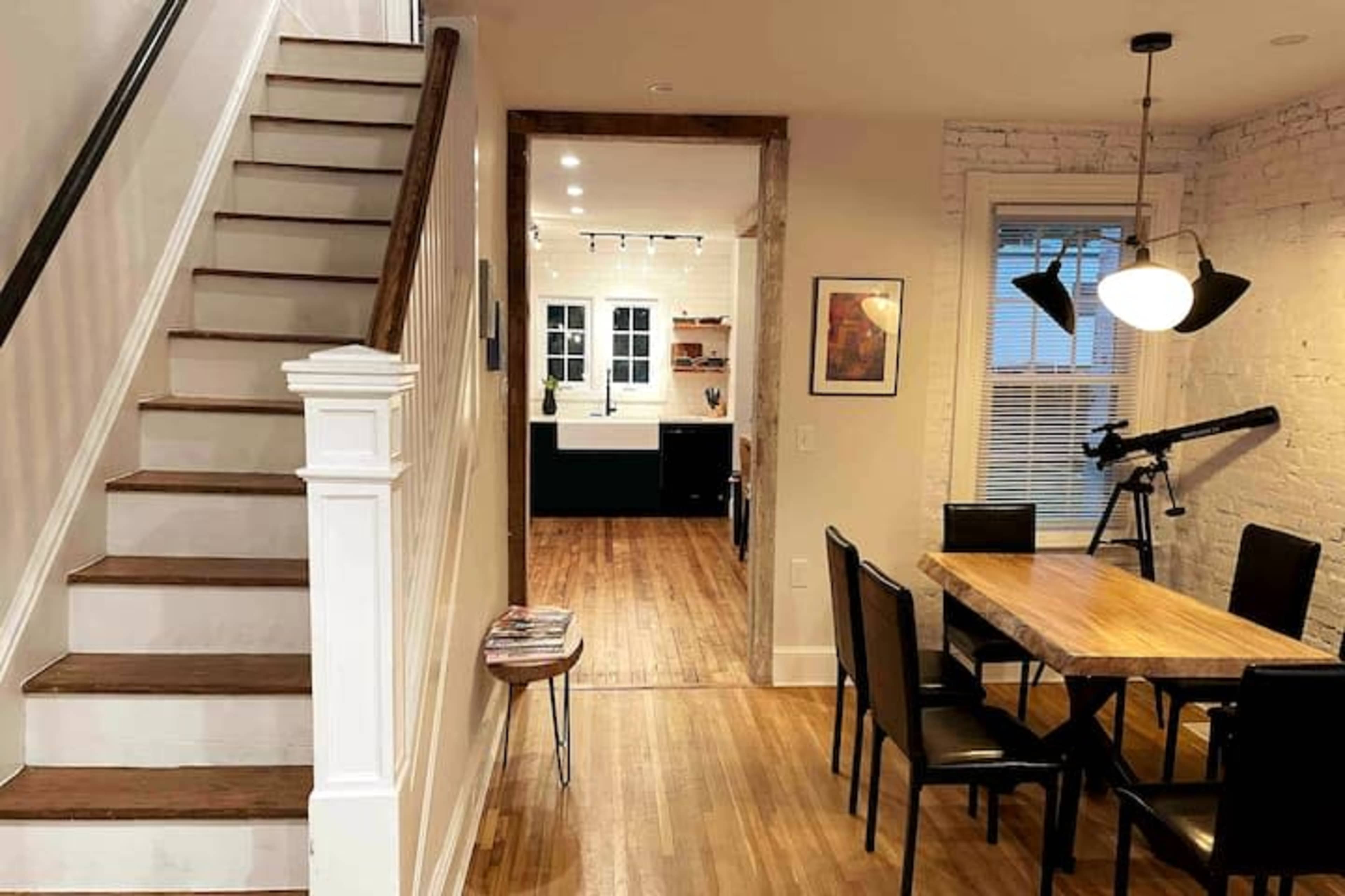 A staircase ascends from a dining area that features a wooden table and black chairs, leading to a kitchen visible through an open doorway.