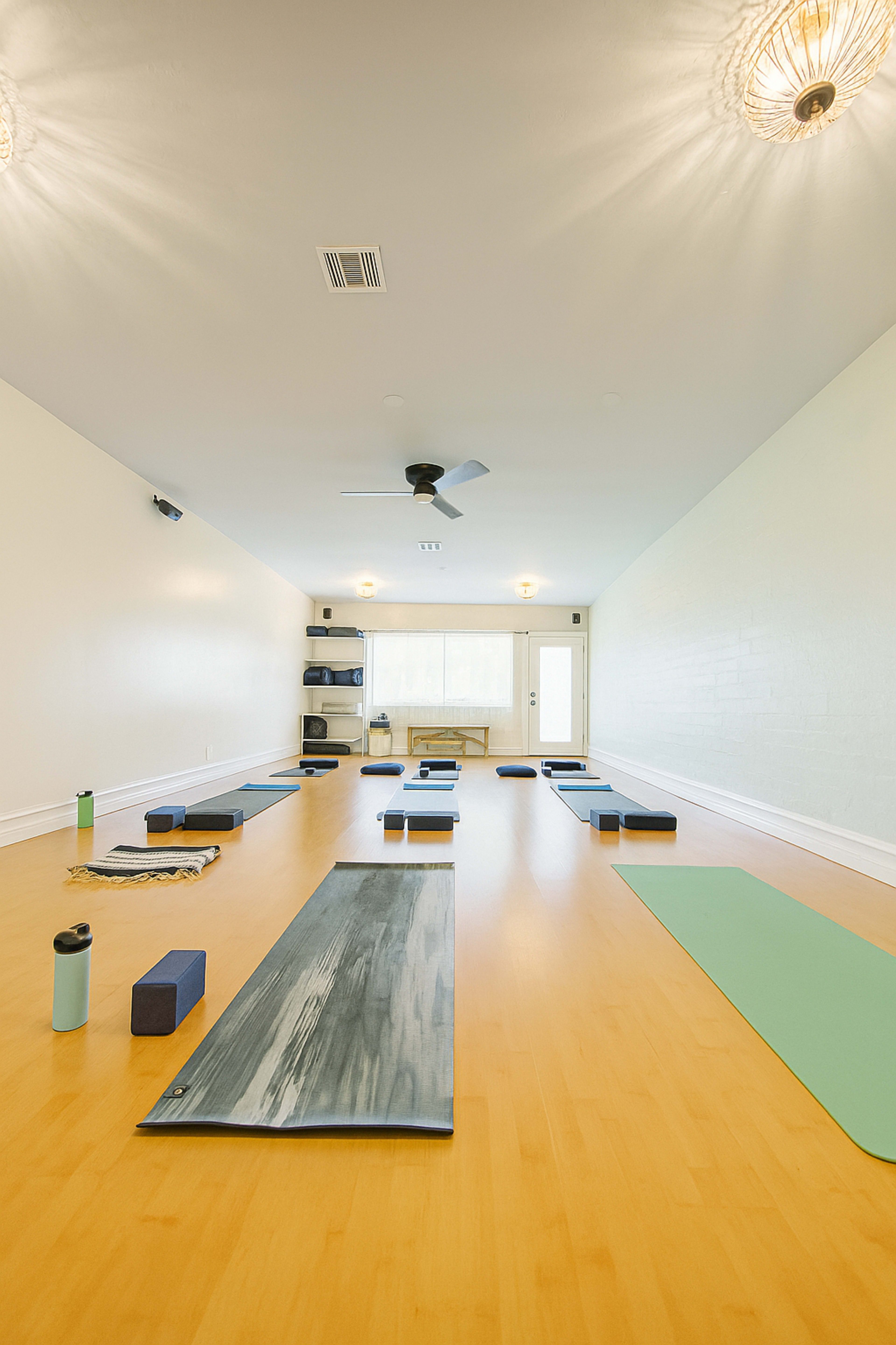 The image shows a spacious, well-lit yoga studio with several yoga mats arranged neatly on a wooden floor, and props like blocks and water bottles placed nearby.