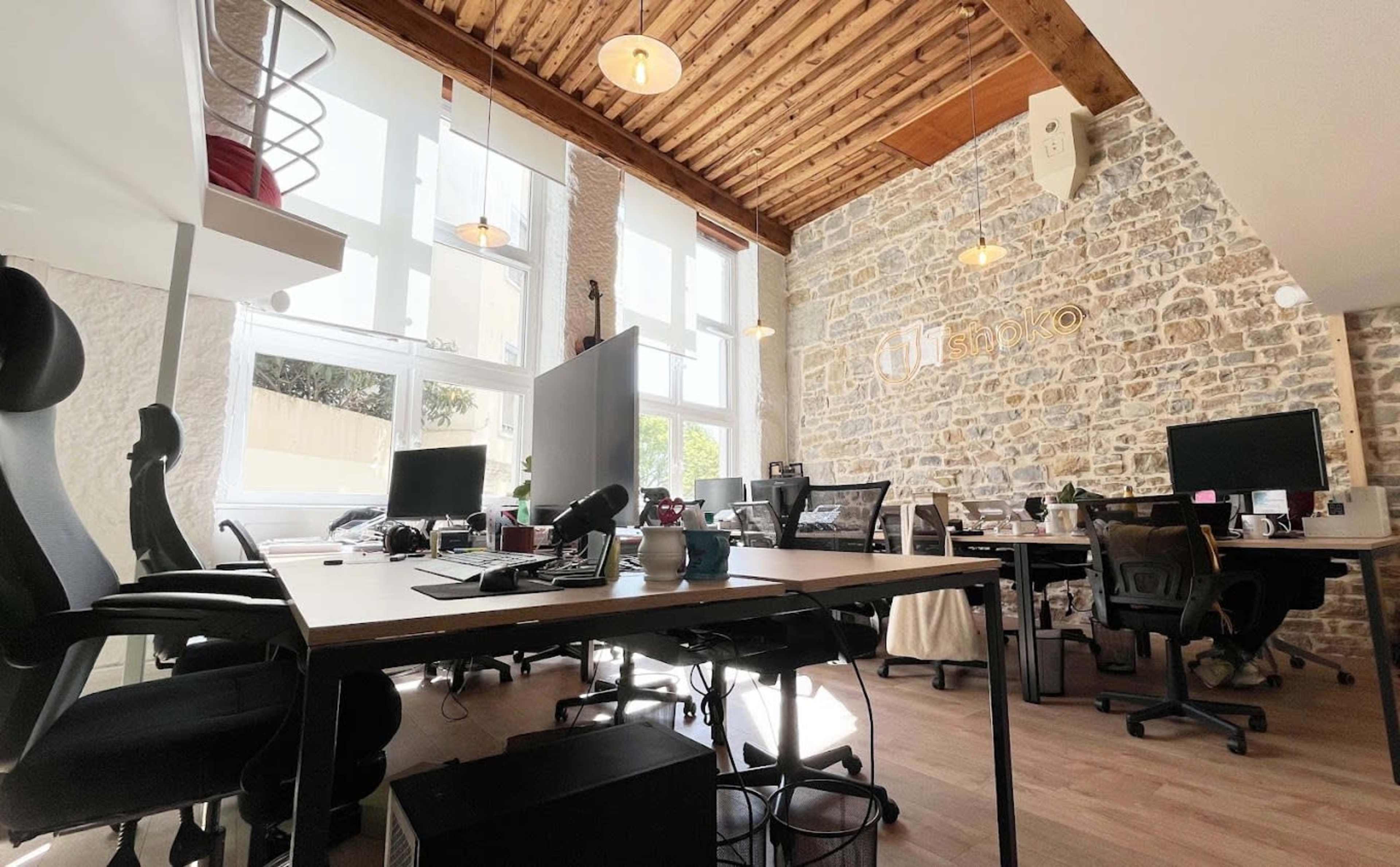 The image shows a modern office space with several work desks, computer monitors, and a mix of ergonomic chairs against a backdrop of exposed stone walls and wooden ceiling beams.