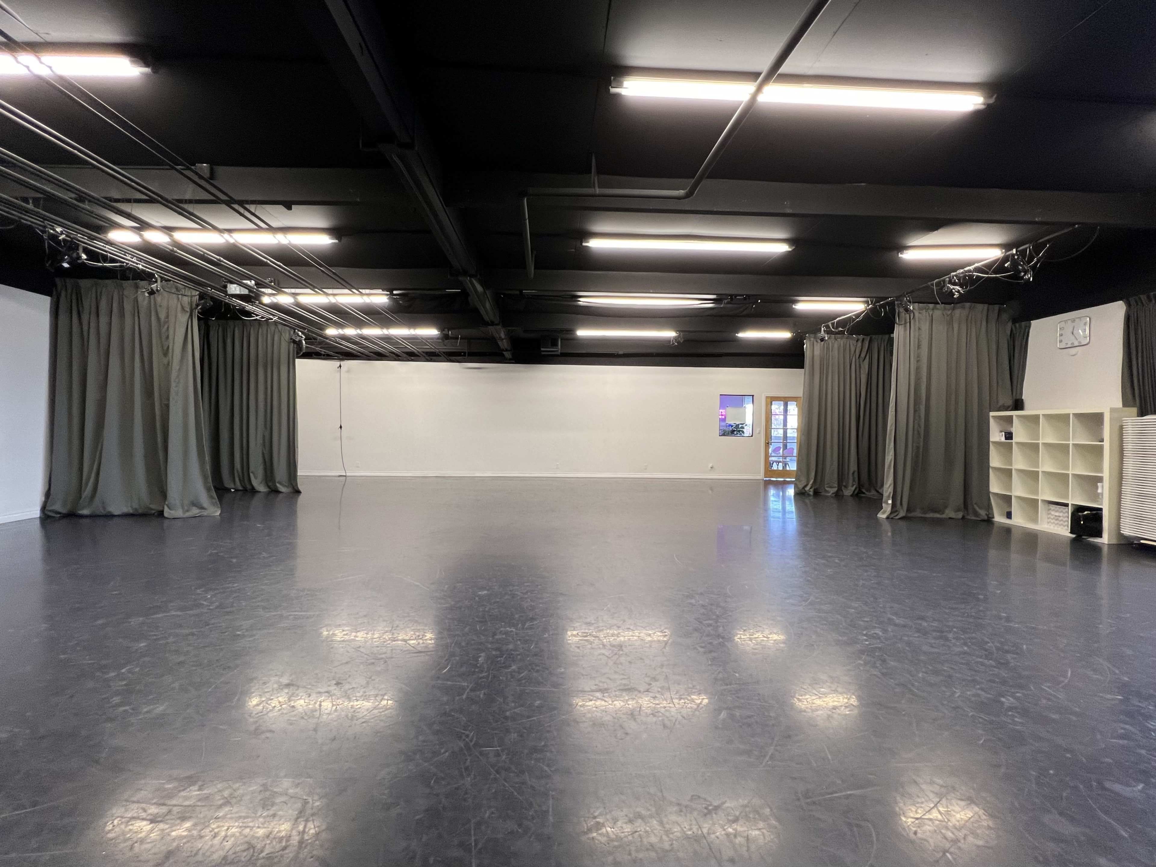 The image shows a spacious dance studio with black flooring, gray curtains, and bright overhead lights.