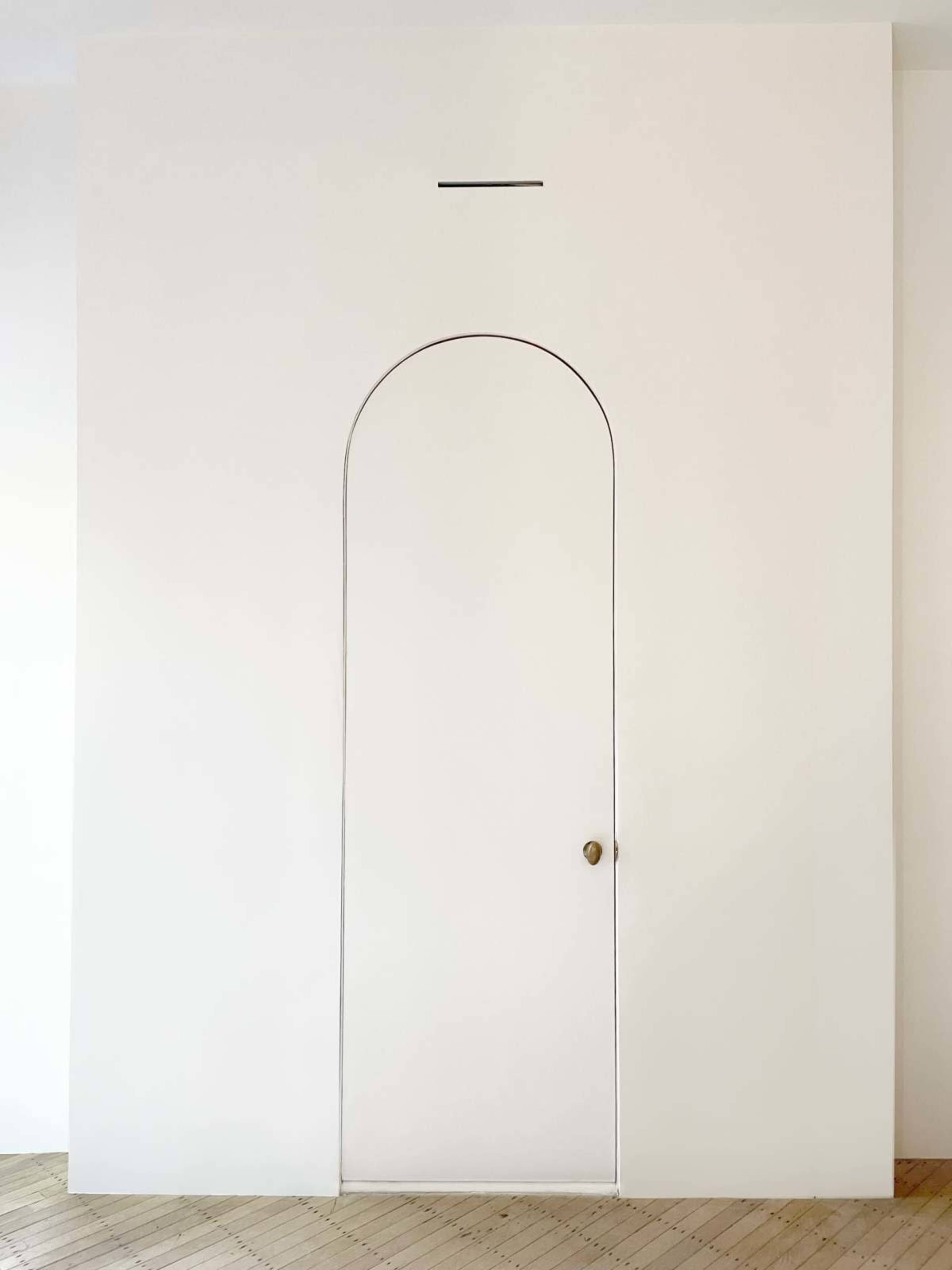 The image shows a plain white wall with an arched doorframe that has a small rectangular slot above it.