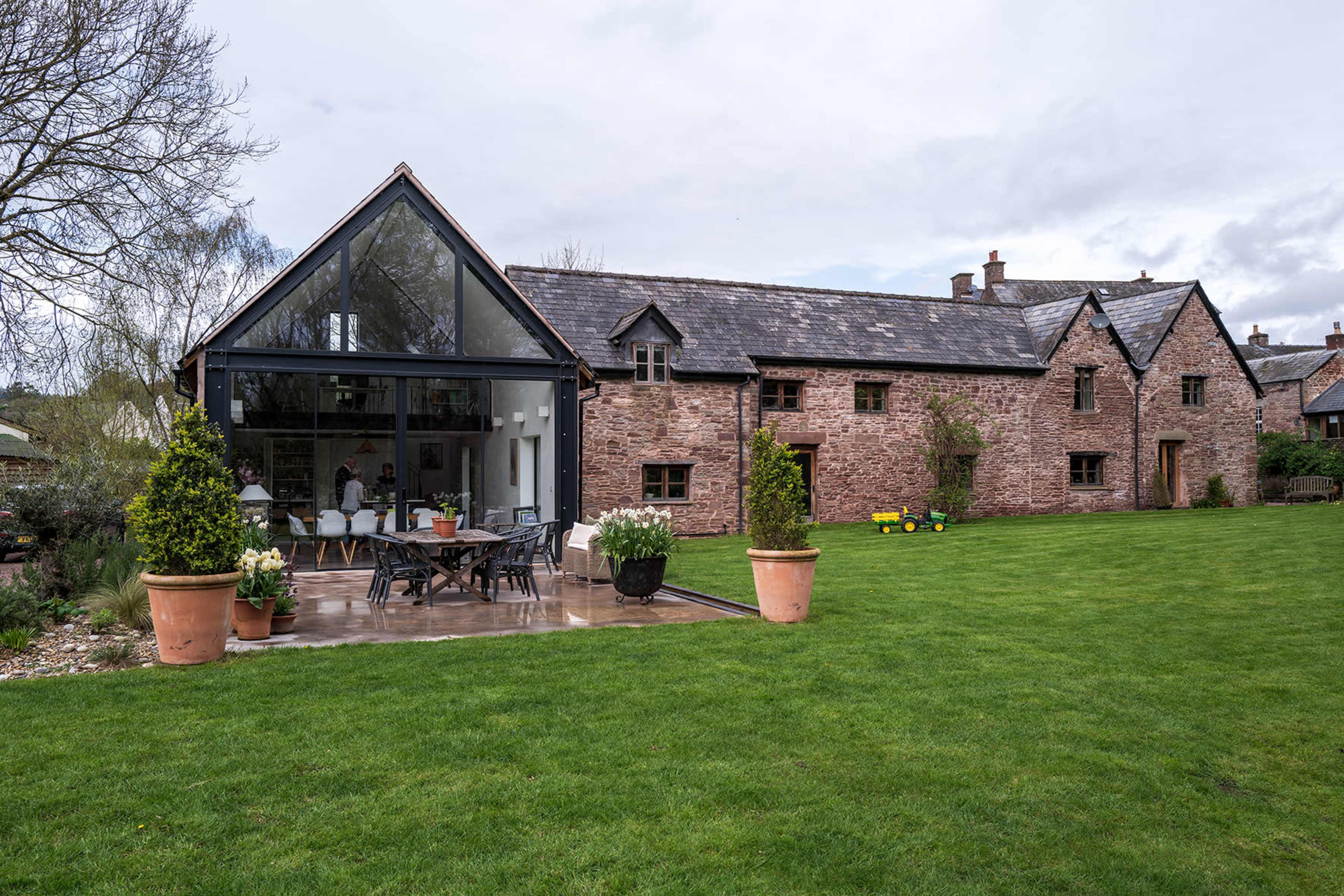 A modern glass extension adjoins a rustic stone farmhouse surrounded by a well-maintained lawn and garden.