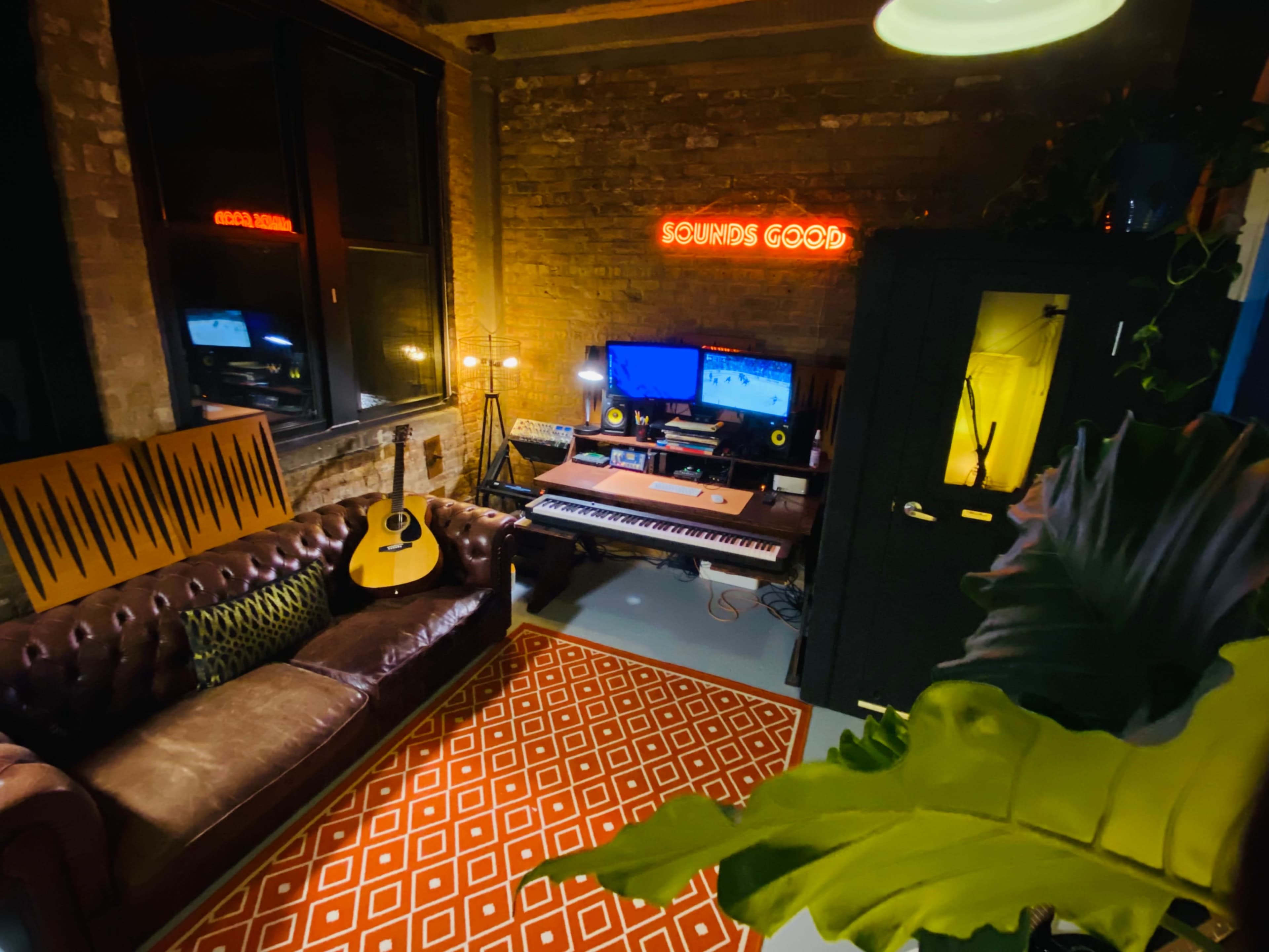 The image shows a cozy music production space featuring a brown leather couch, a keyboard and computer setup, an acoustic guitar, and a neon sign that reads "SOUNDS GOOD."