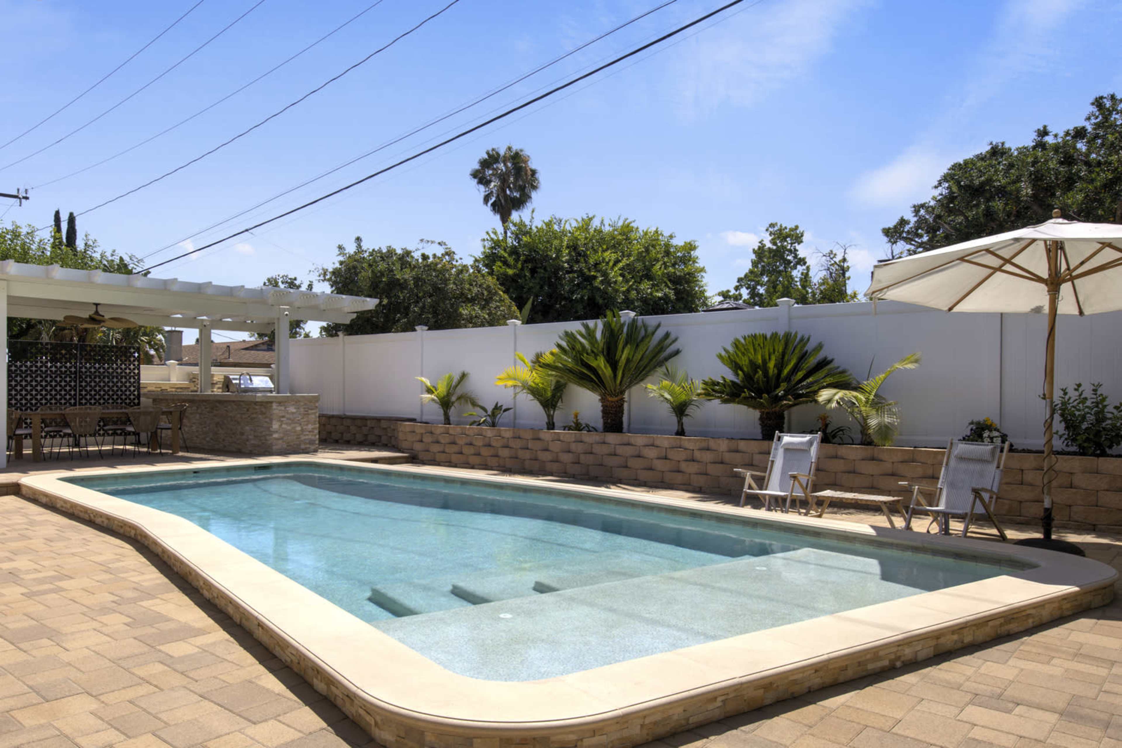 The image shows a rectangular swimming pool surrounded by a patio, palm trees, and outdoor furniture beneath an umbrella.