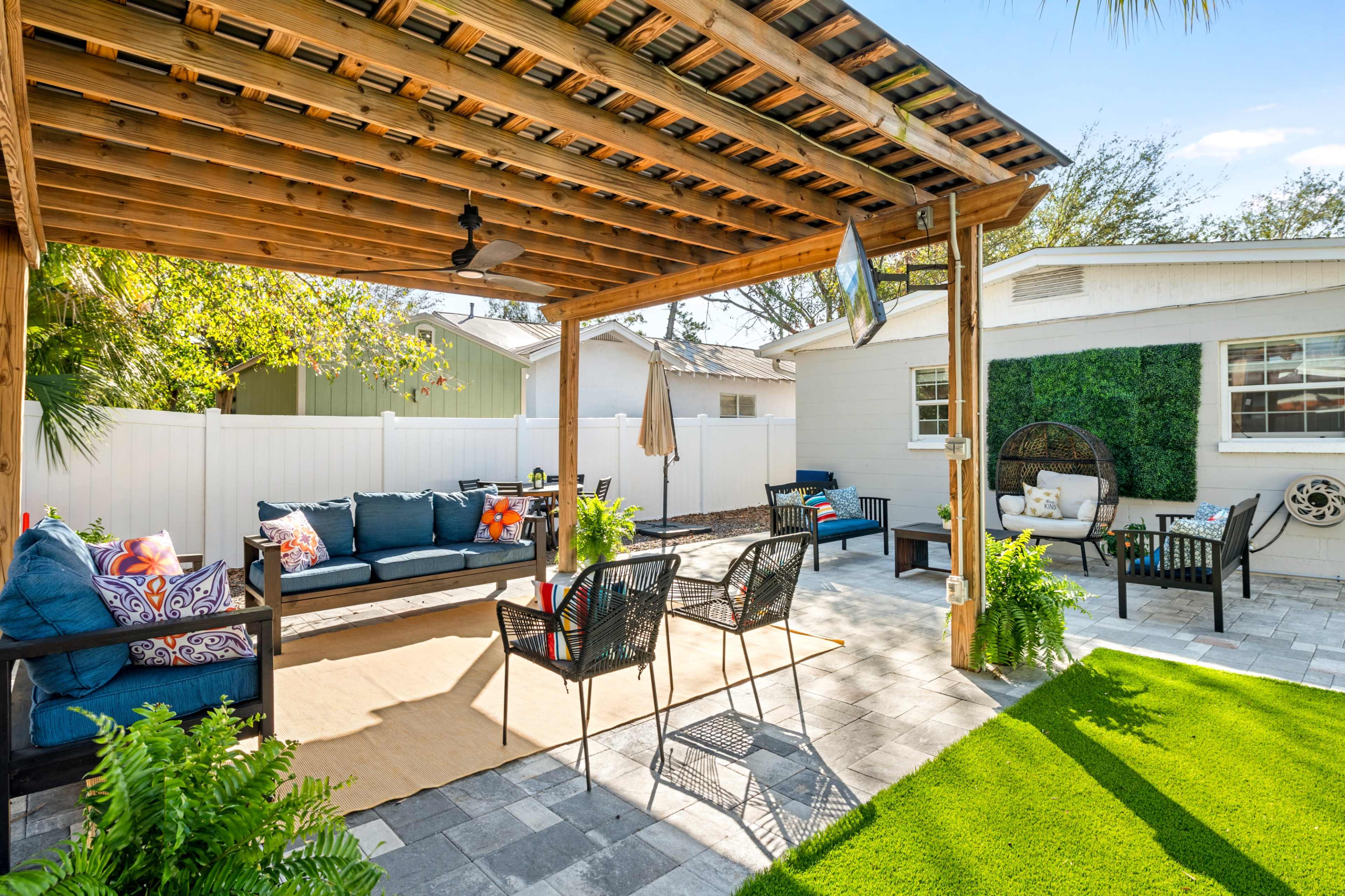 The image shows a backyard patio with a wooden pergola, seating areas featuring sofas and chairs, and a landscaped green area with decorative plants.