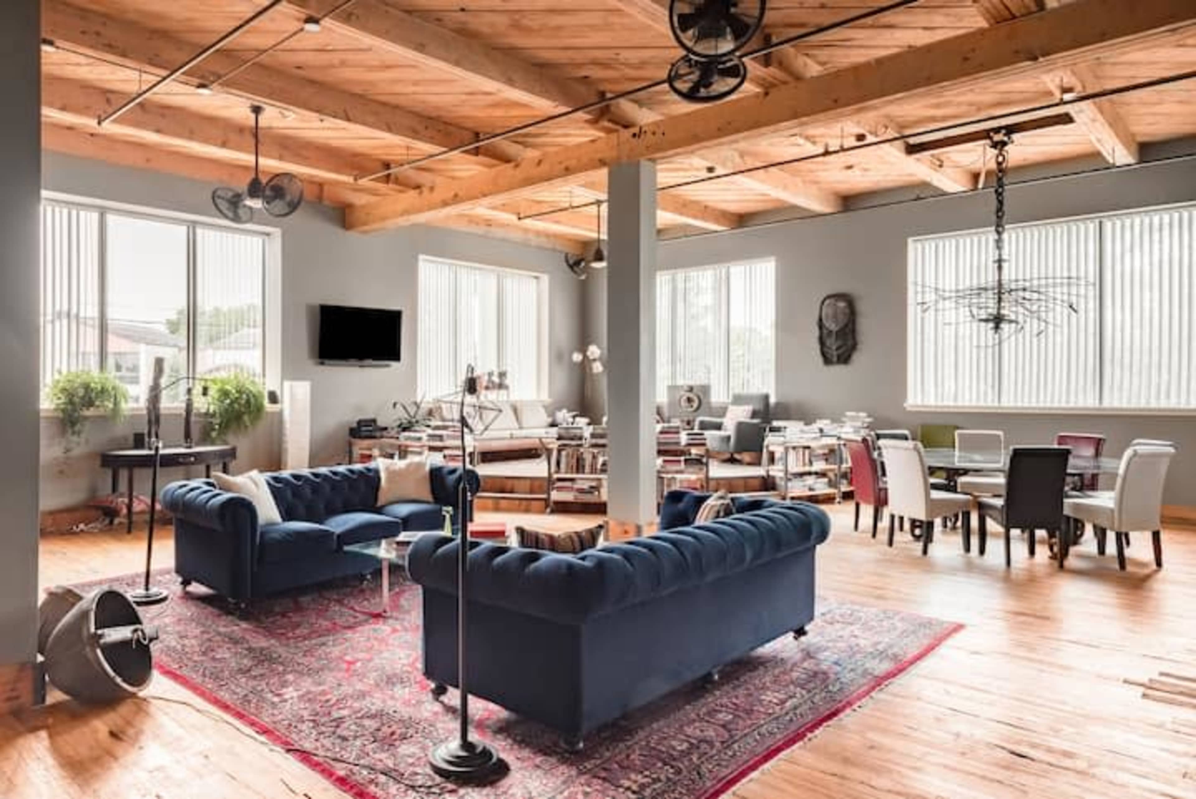A spacious, modern open-concept living area with wooden beams, a large area rug, and various seating arrangements near numerous windows.