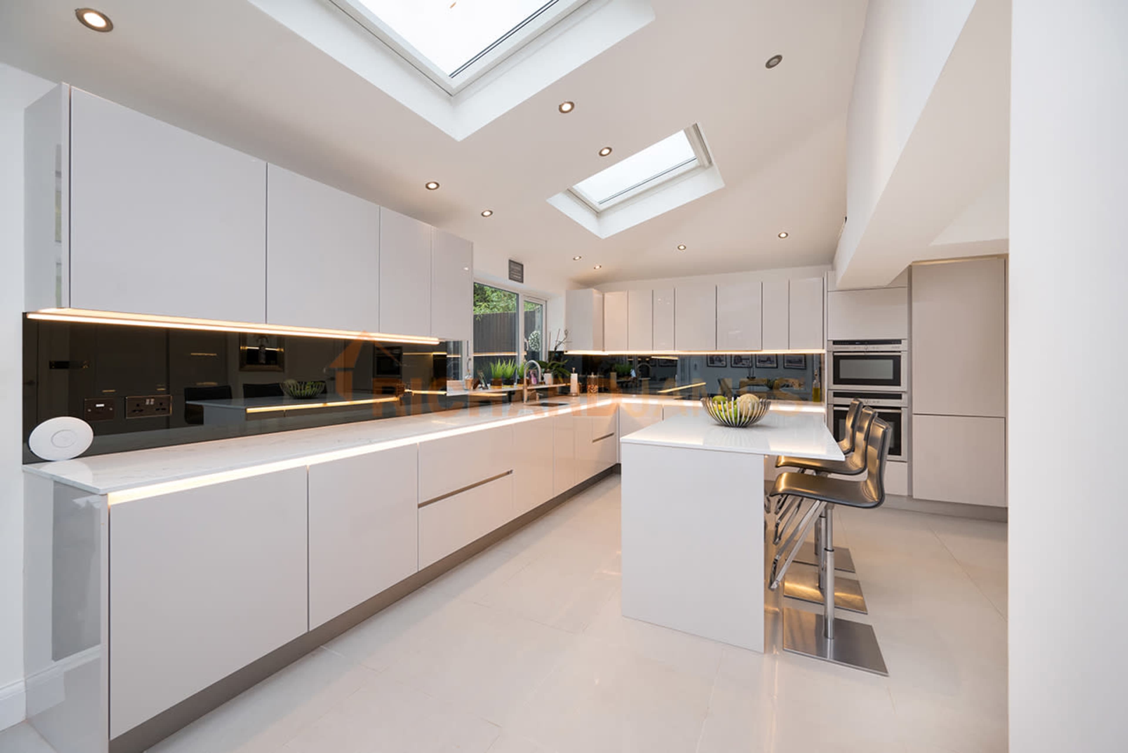 A modern kitchen with sleek white cabinetry, a central island with bar stools, and large skylights letting in natural light.