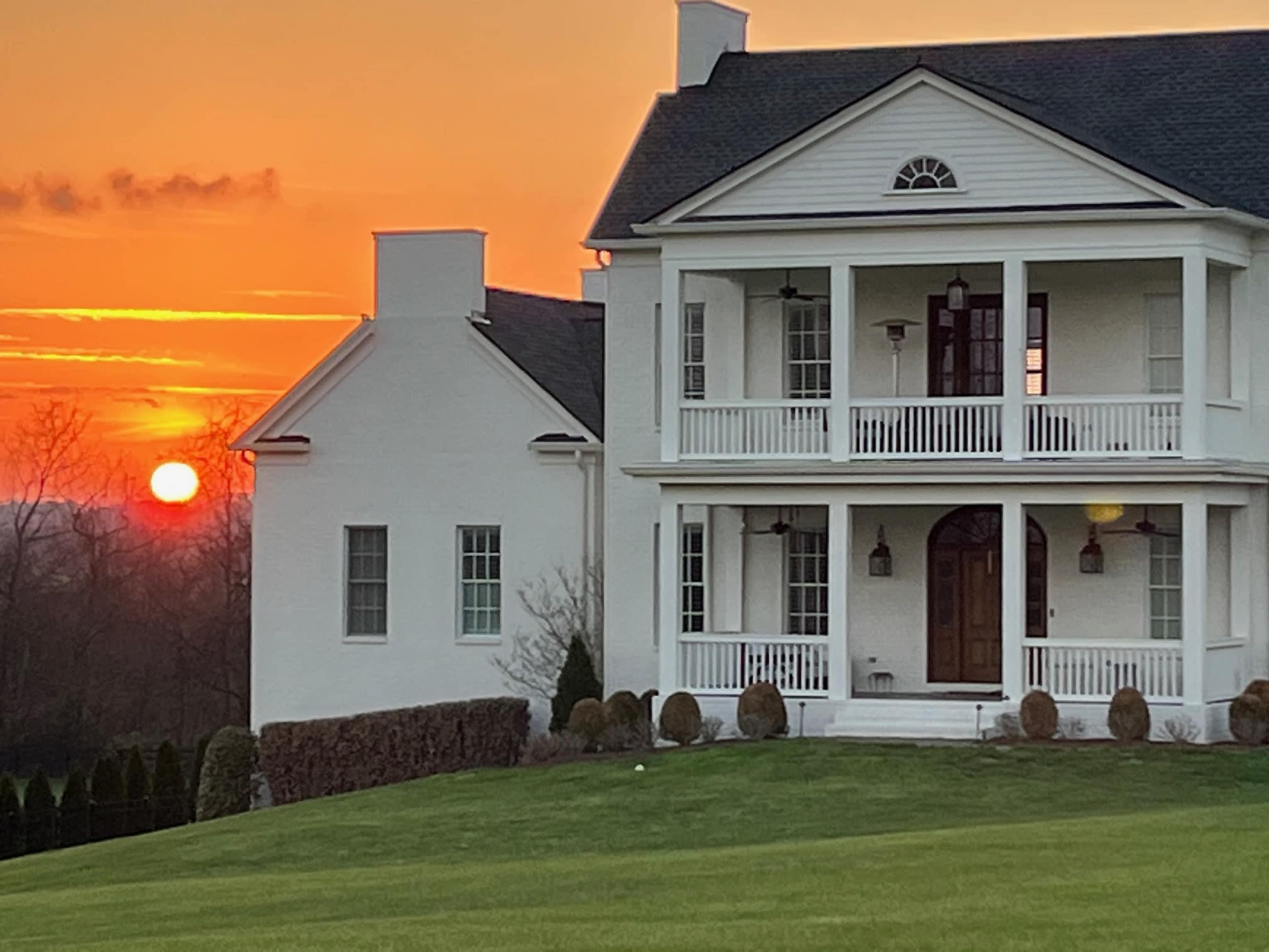 A white house with multiple balconies stands against a backdrop of a sunset, highlighting the sun partially obscured by the horizon.