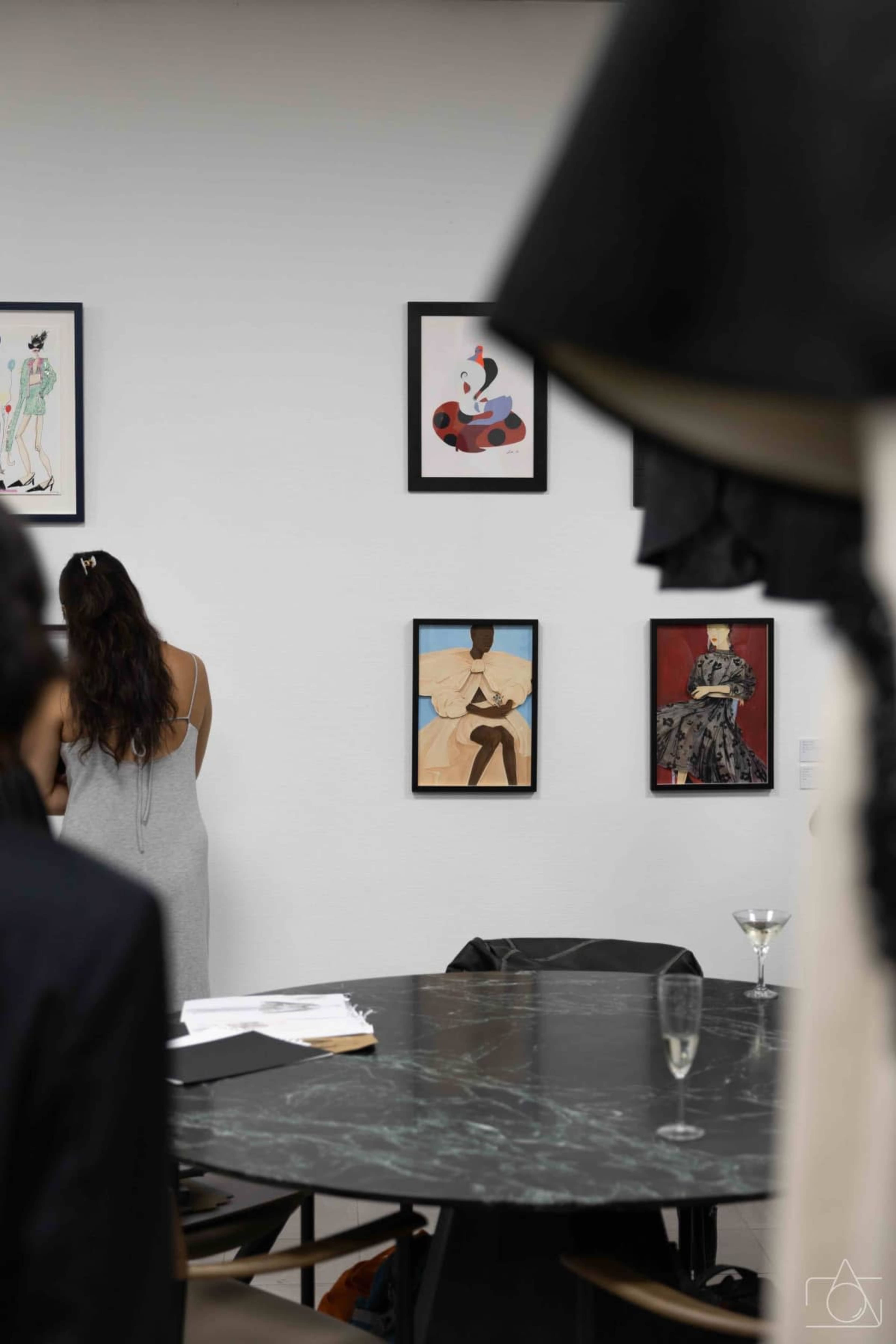 A person stands facing a wall adorned with framed artwork in an art gallery, while a table with a dark marble surface is visible in the foreground.