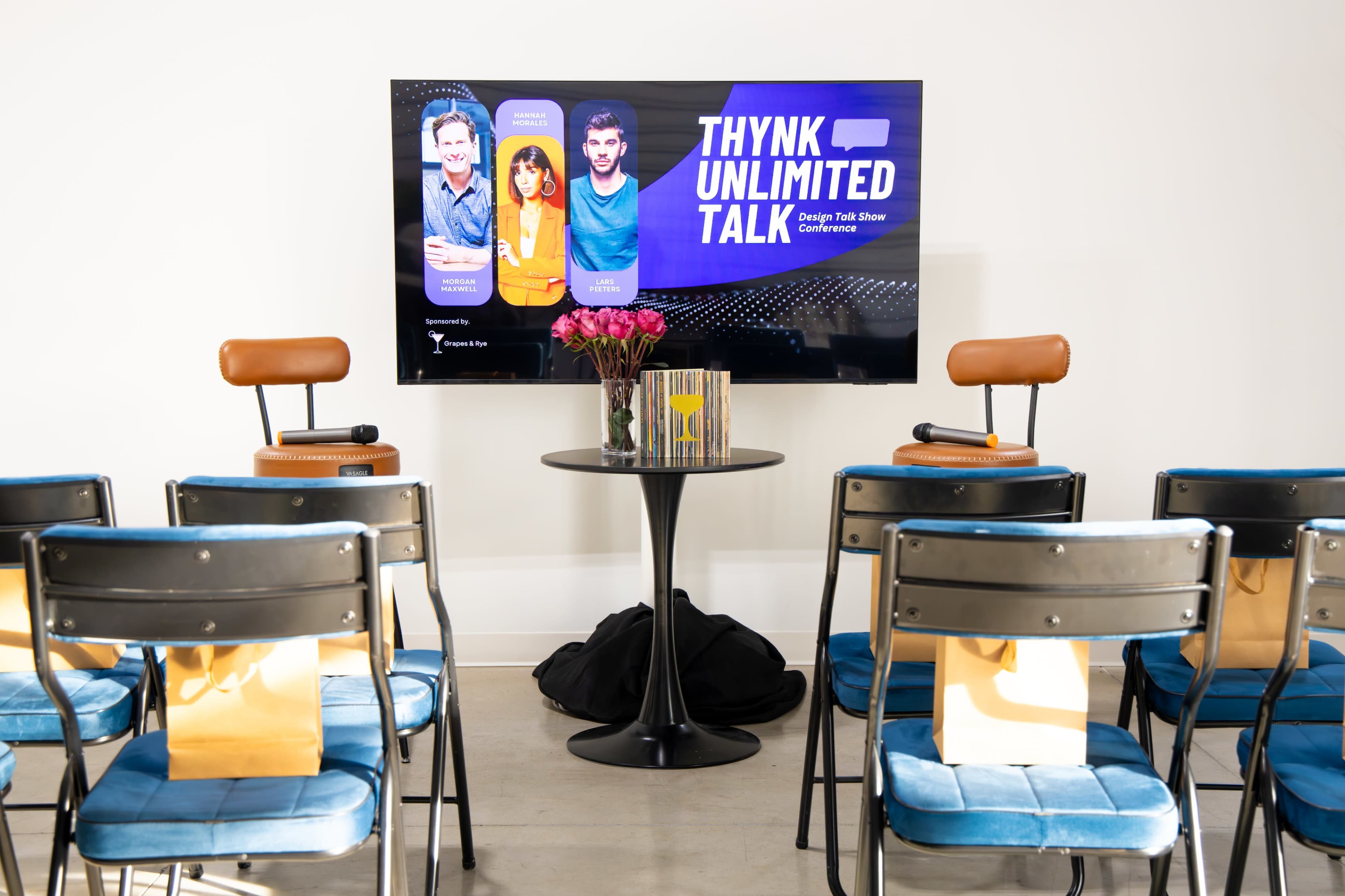 The image shows a conference setup with a large screen displaying "THYNK UNLIMITED TALK," surrounded by empty chairs and a small table with a vase of flowers.
