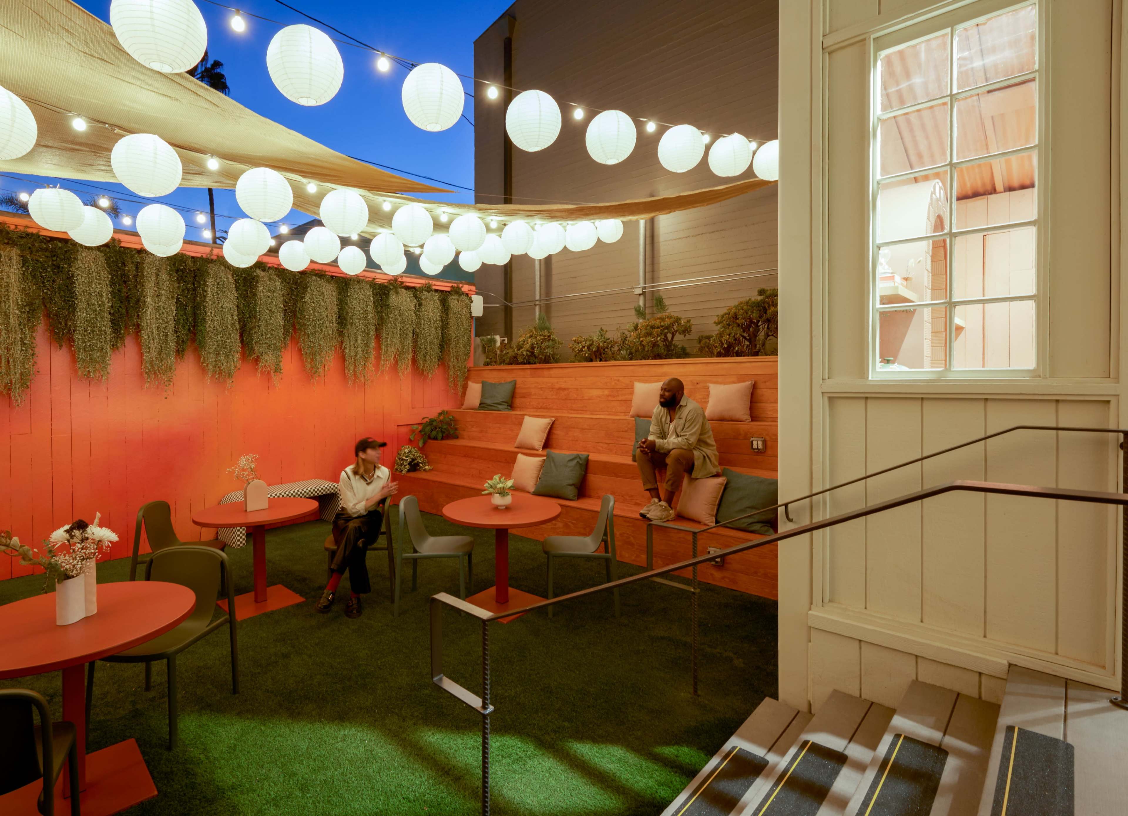 An outdoor seating area with circular tables, green chairs, and decorative string lights above, set against a backdrop of a vibrant orange wall and greenery.
