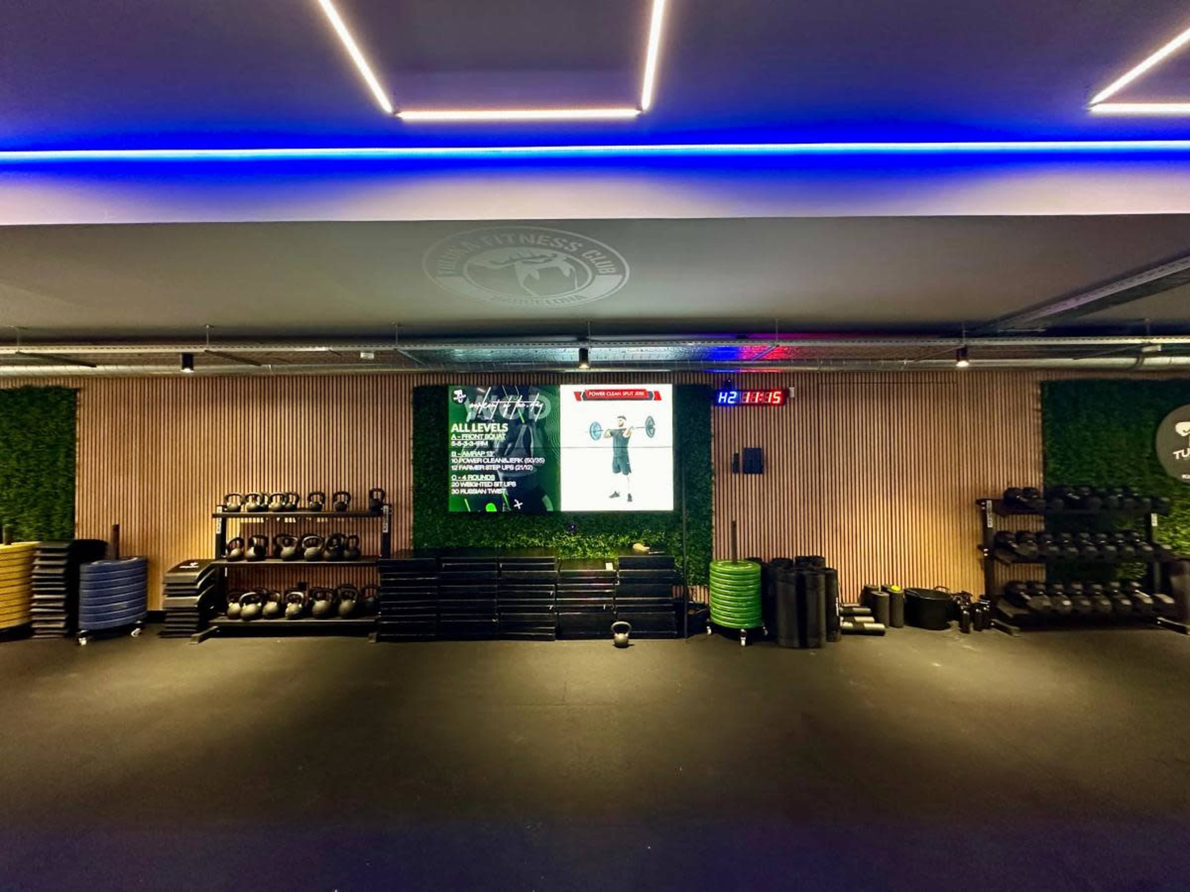 A modern gym interior with organized workout equipment, a large screen displaying fitness information, and green wall accents.
