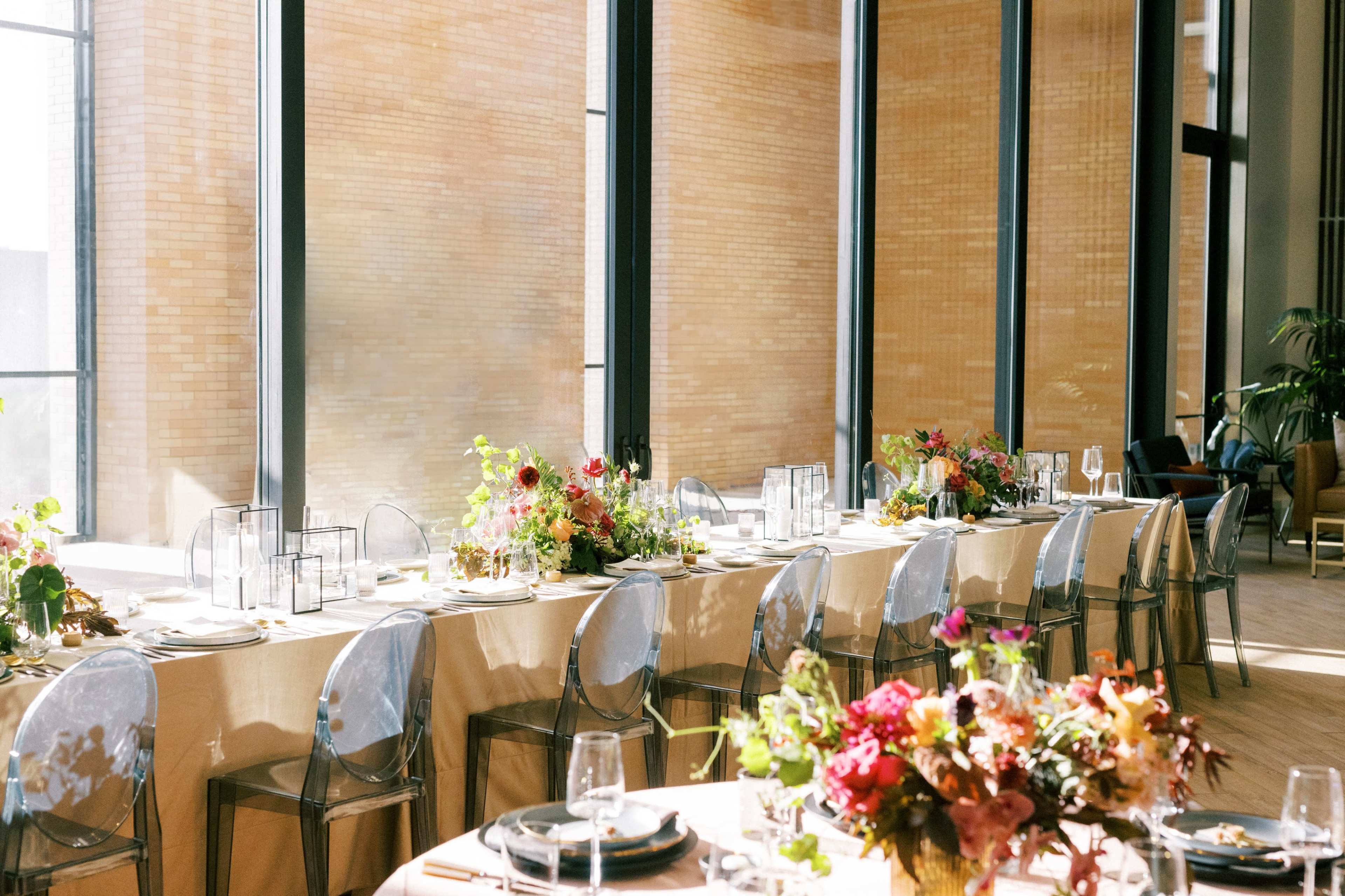 A long dining table is set with floral arrangements and glassware, illuminated by natural light from large windows.