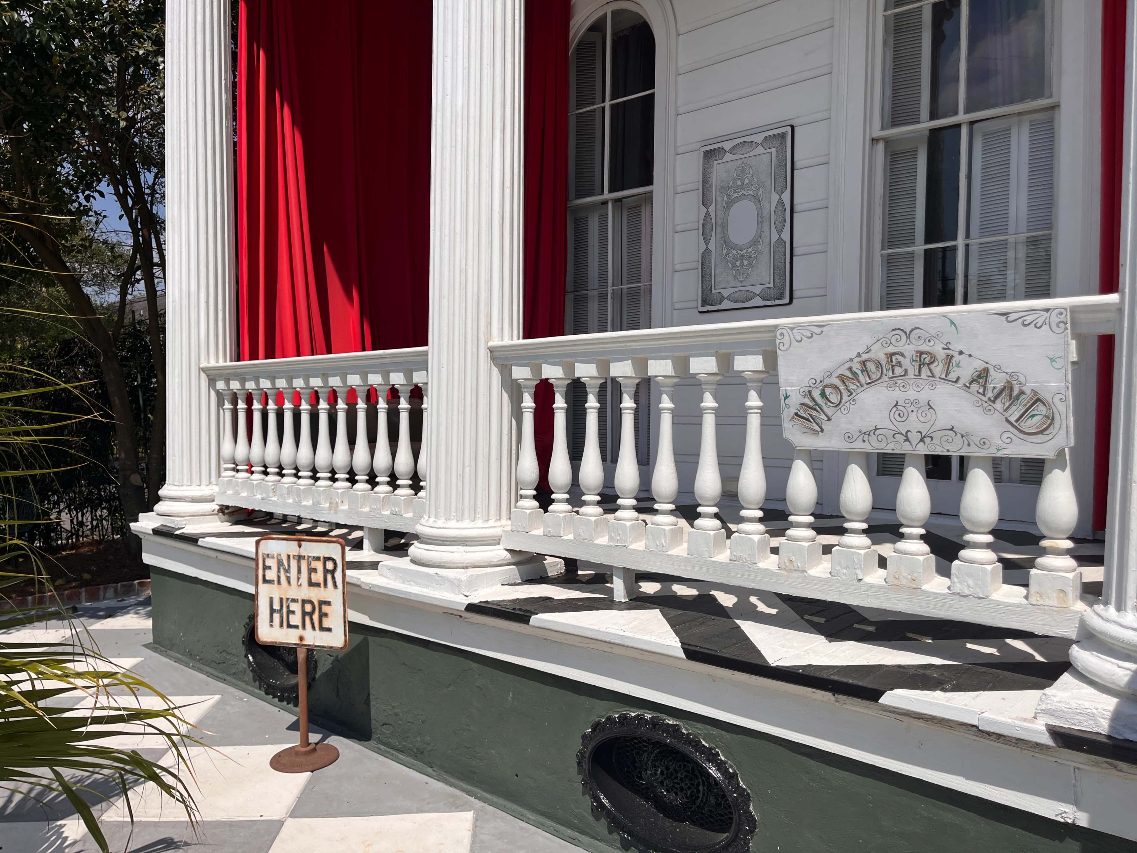 The image shows a porch with white columns, a sign reading "WONDERLAND," and a wooden sign that says "ENTER HERE" placed on the ground.