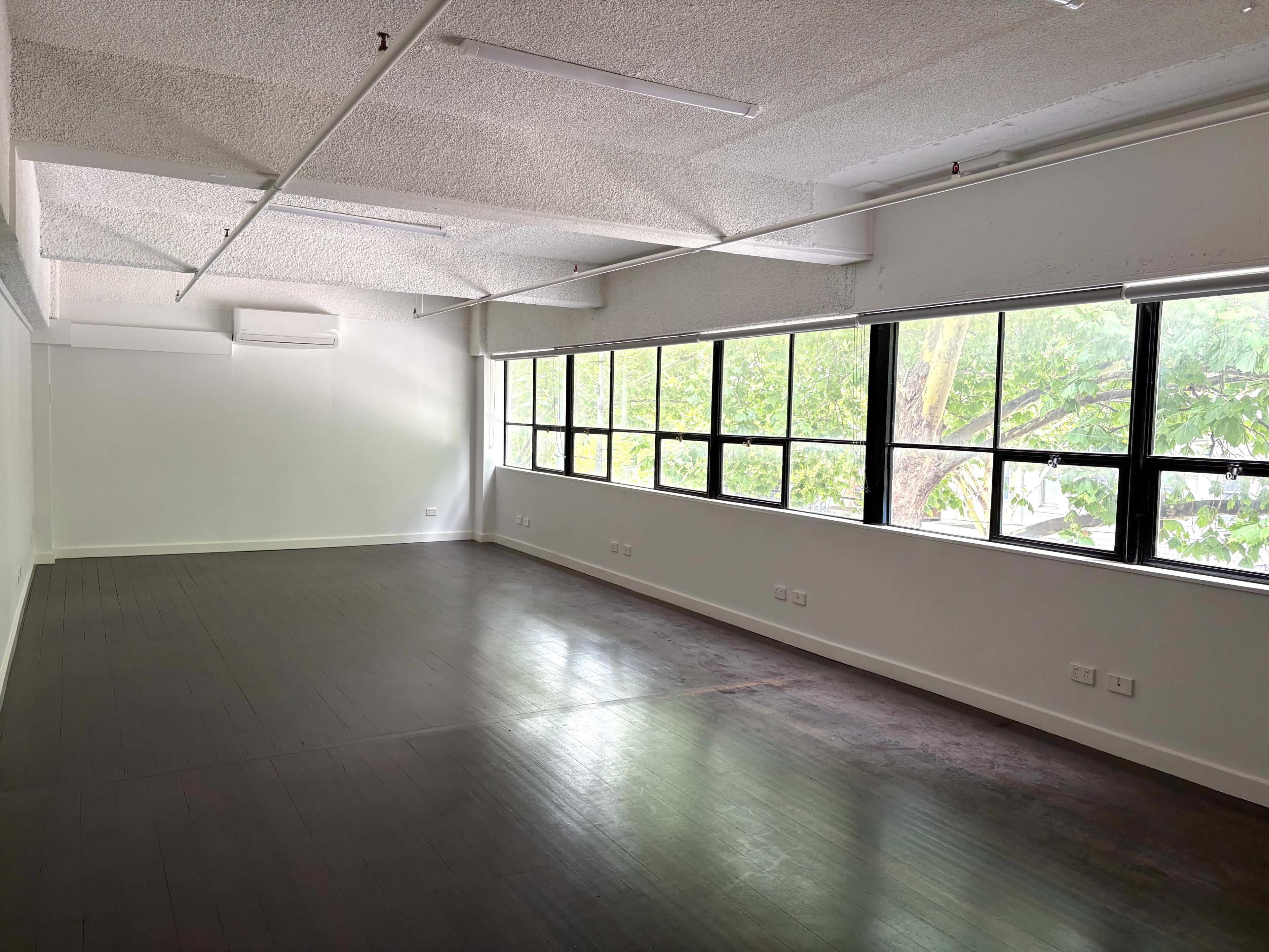 The image shows an empty interior space with large windows allowing natural light to enter, featuring a smooth wooden floor and plain white walls.