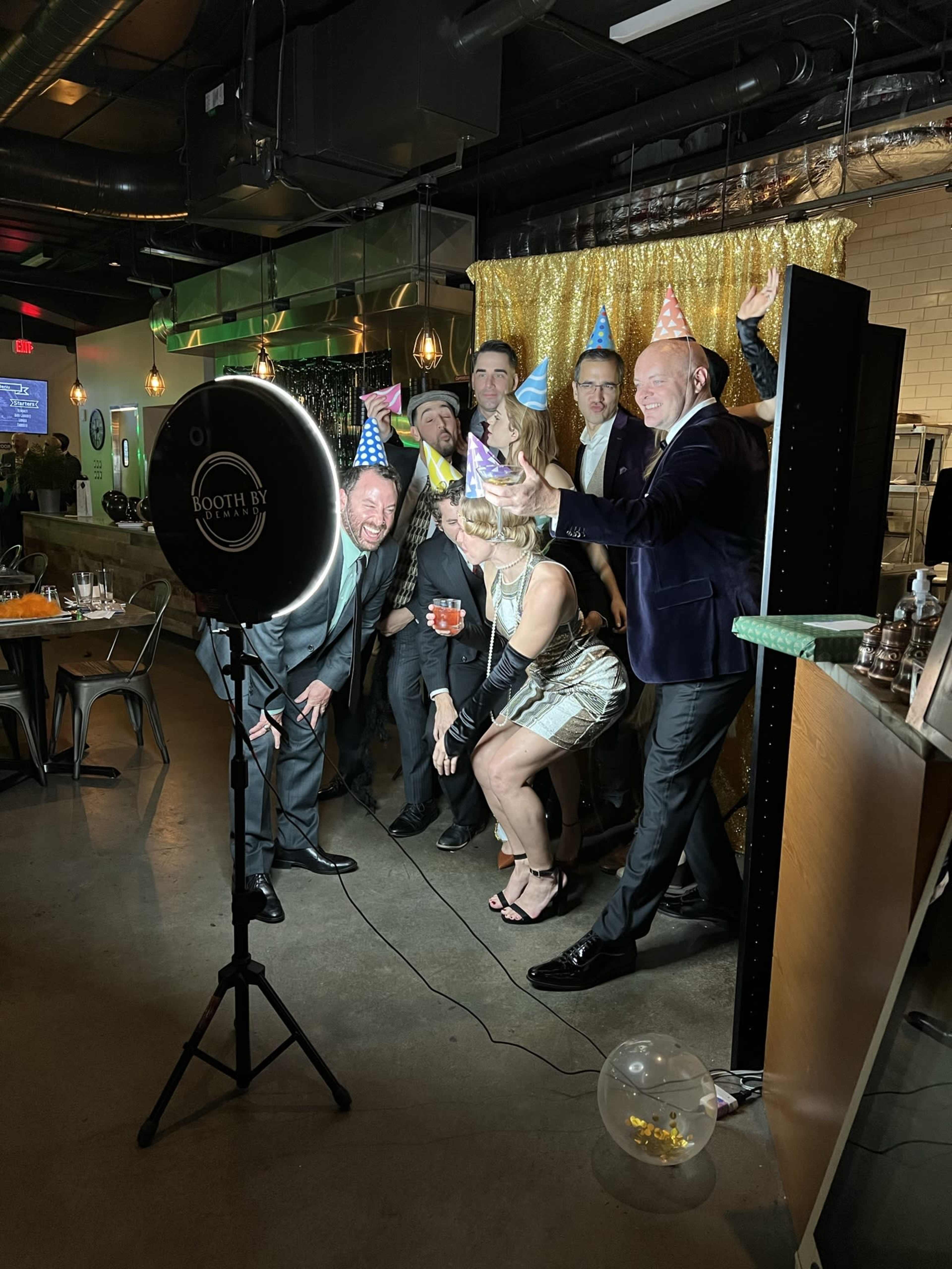 A group of seven people wearing party hats poses for a photo in front of a sparkling gold backdrop.