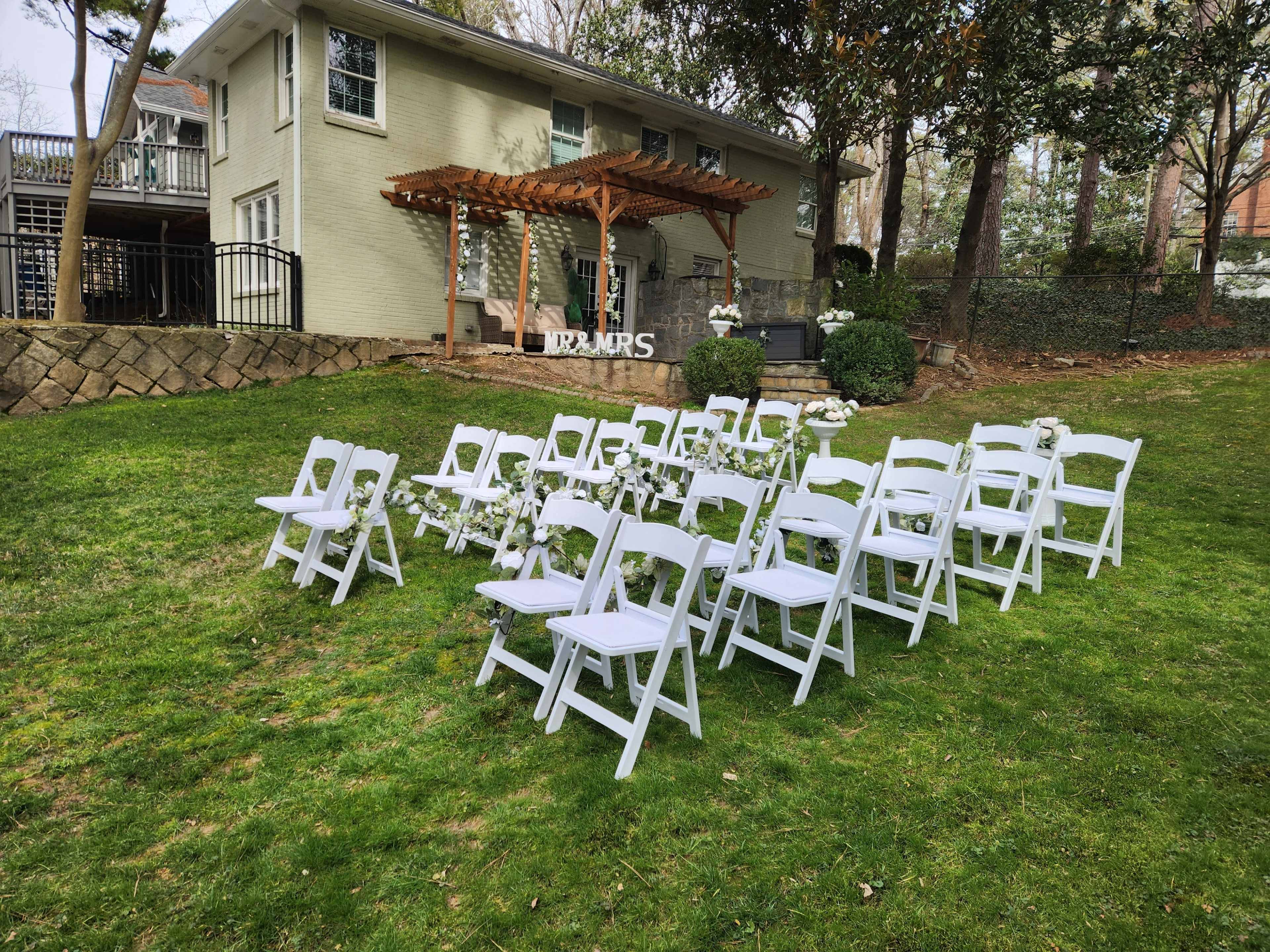 A wedding setup featuring rows of white folding chairs arranged on a green lawn in front of a house with a wooden pergola.