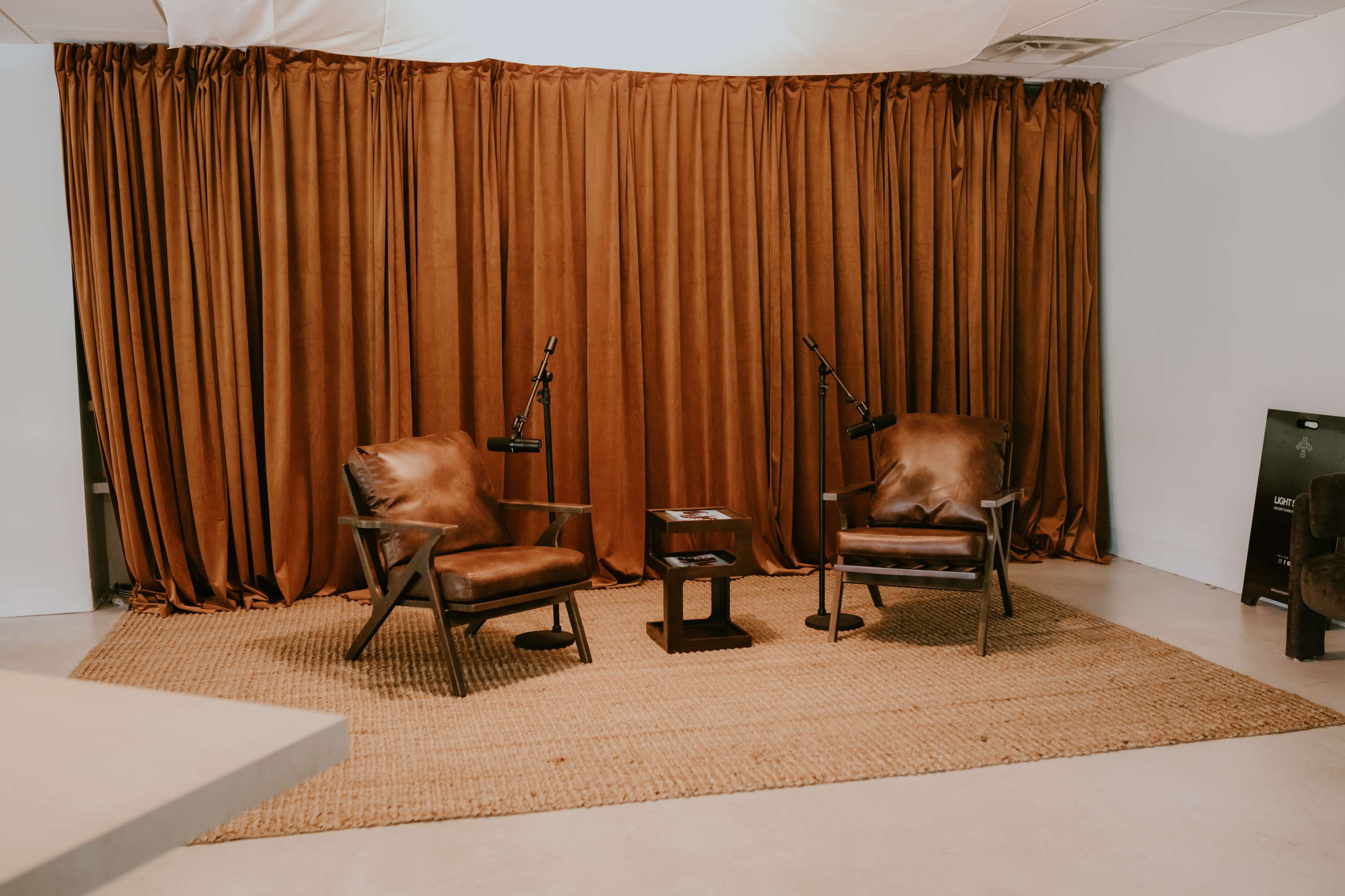 The image shows two brown leather armchairs with floor lamps on either side, positioned in front of a large brown curtain and a small table, all set on a woven rug in a minimalist room.