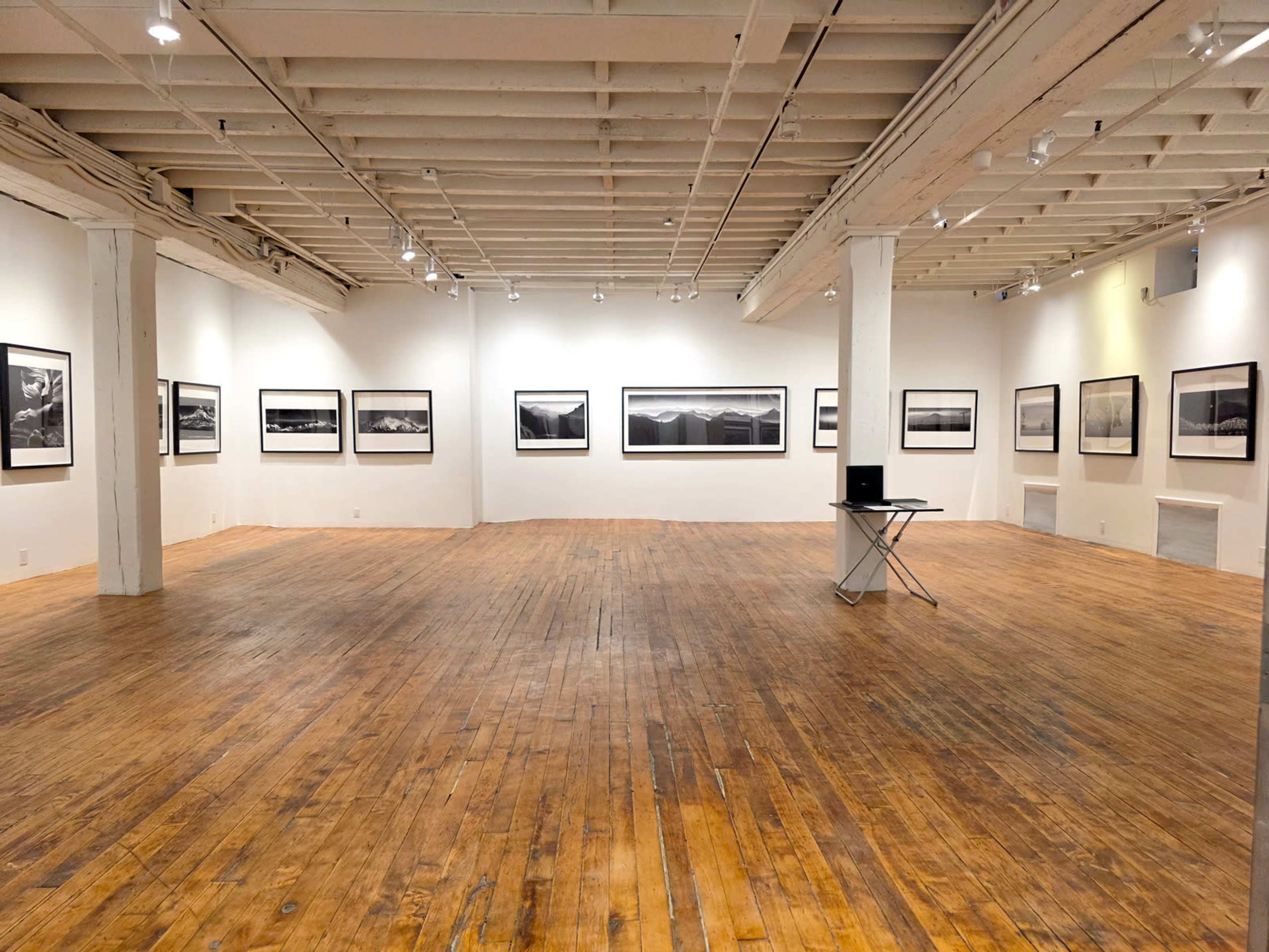 The image shows an empty art gallery with white walls displaying black-and-white photographs and wooden flooring.