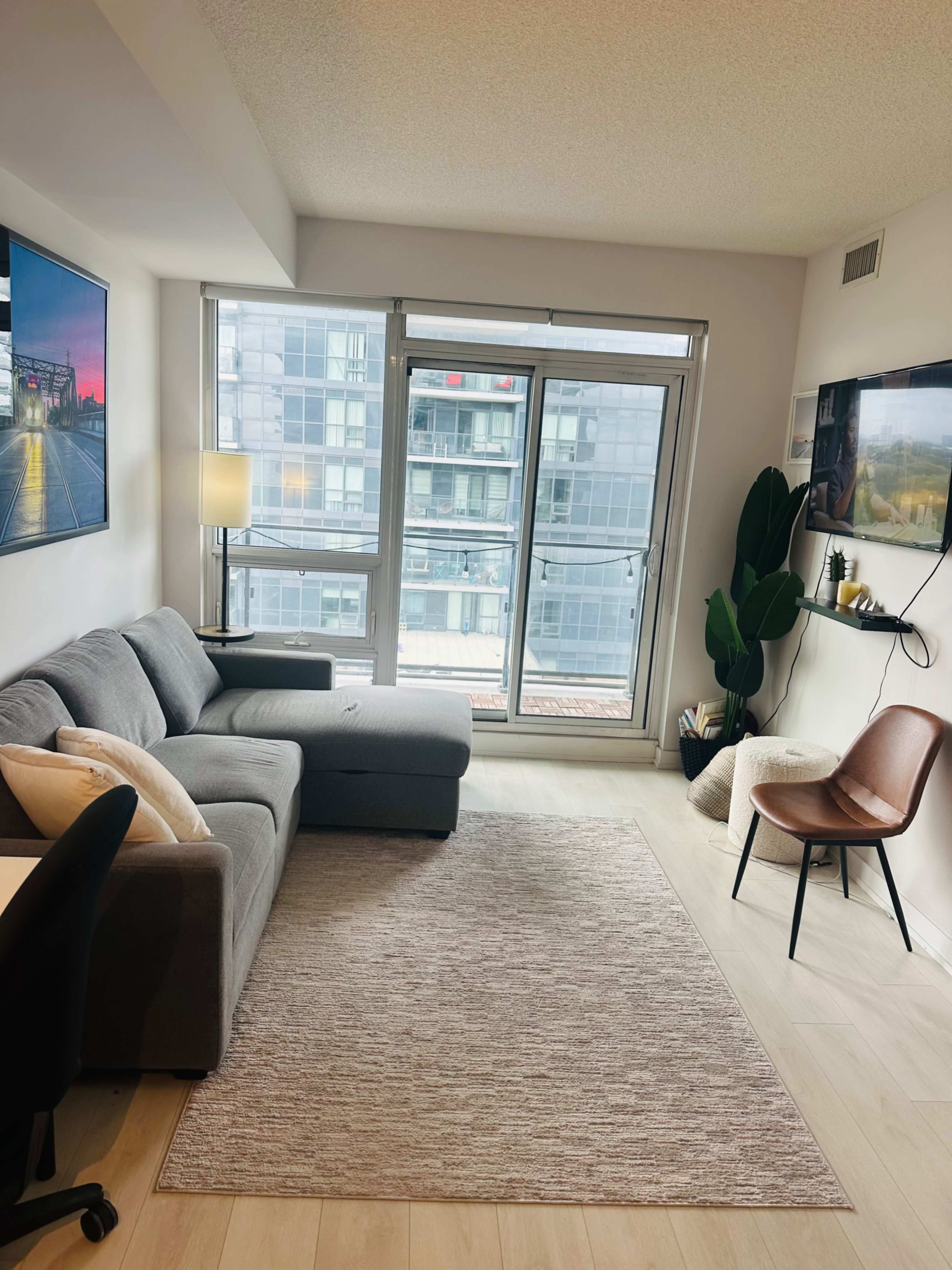 The image shows a modern living room featuring a grey sofa, a large window with city views, a chair, and a small decorative plant.