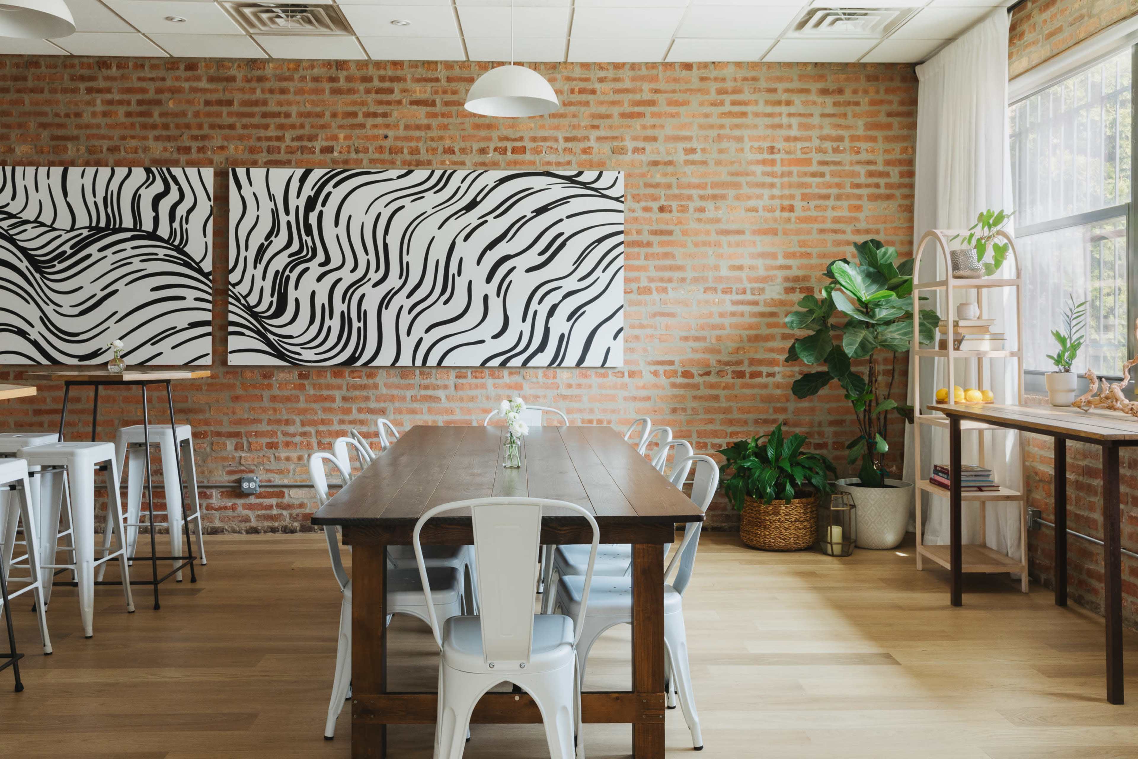 The image shows a dining area with a long wooden table surrounded by metal chairs, a brick wall adorned with abstract black and white artwork, and potted plants in the corners.