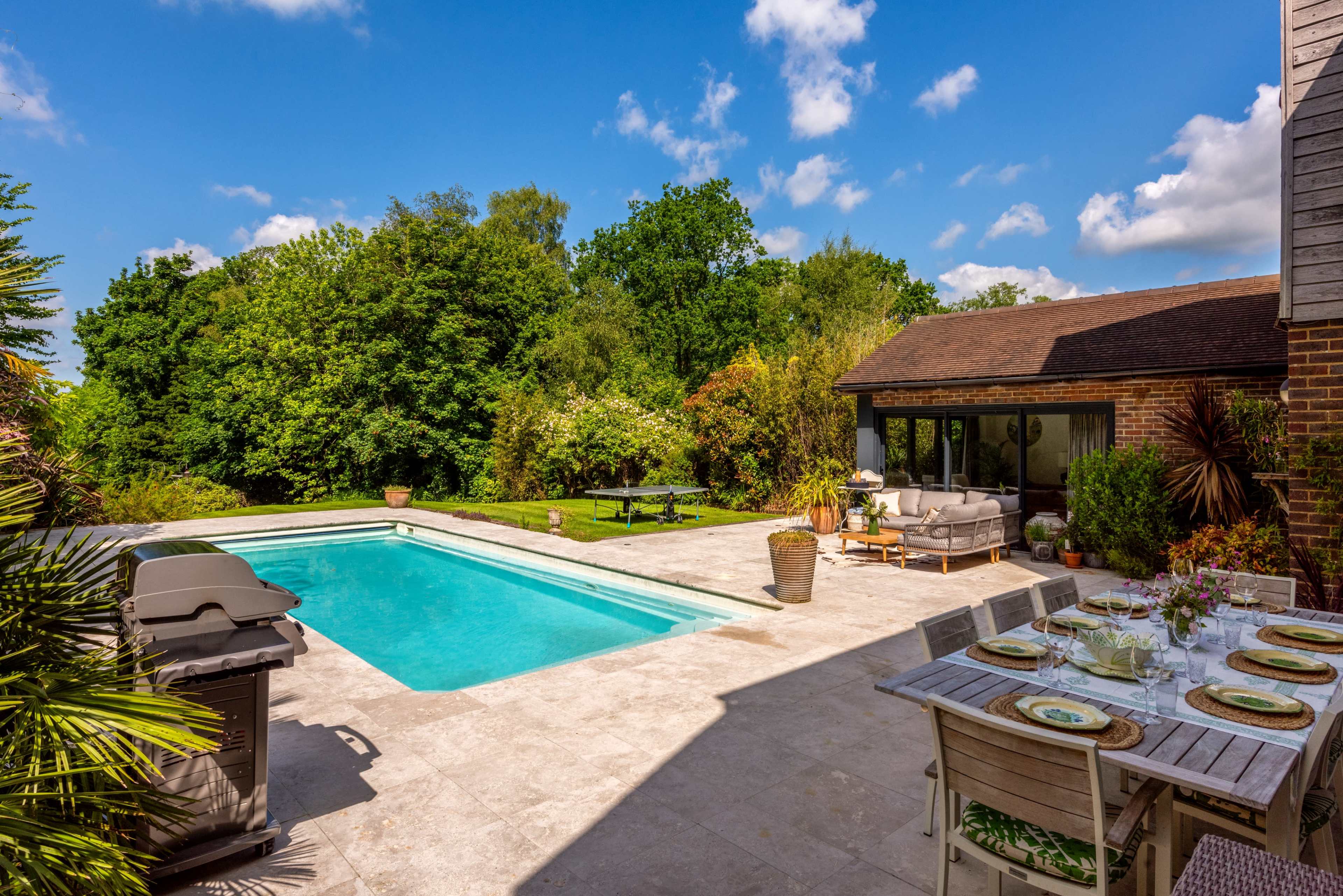 A backyard features a swimming pool surrounded by a patio with outdoor seating and a dining table, set against a backdrop of lush greenery and a clear blue sky.