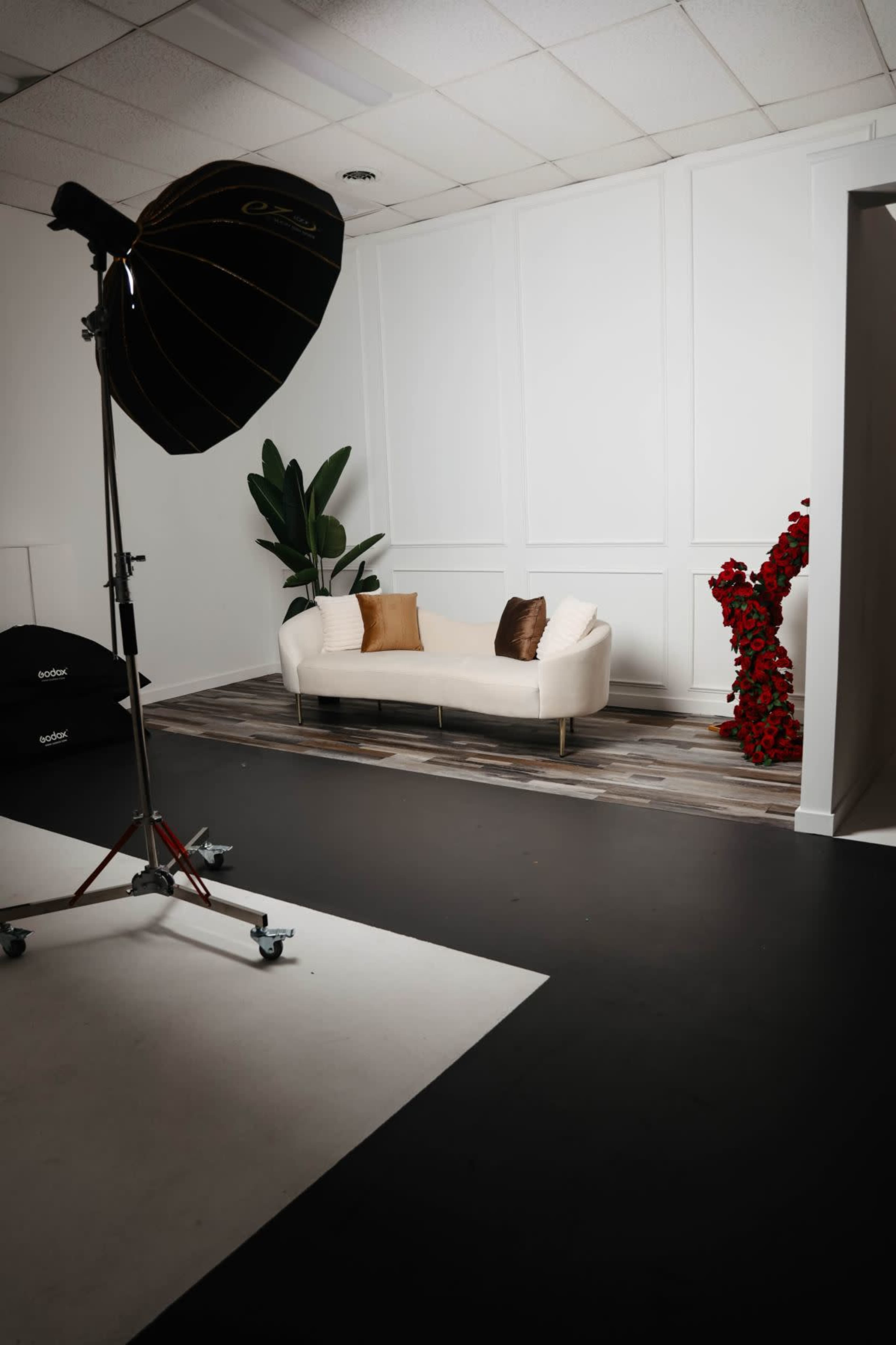 A well-lit photography studio featuring a white couch with two brown pillows, a backdrop of white walls, and a decorative arrangement of red flowers in the corner.