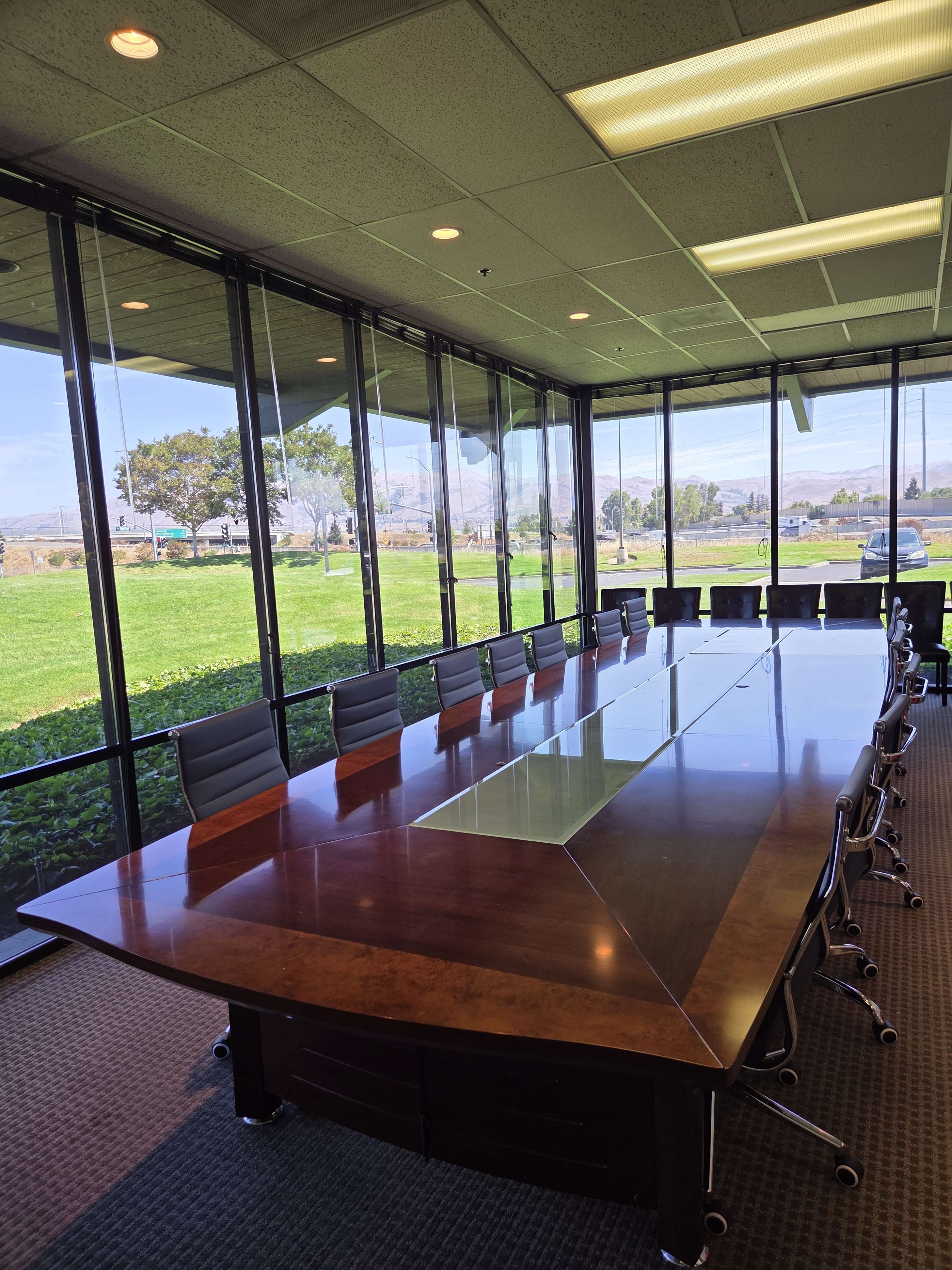 A large conference room features a long wooden table surrounded by black chairs, with floor-to-ceiling windows offering views of green fields and trees outside.