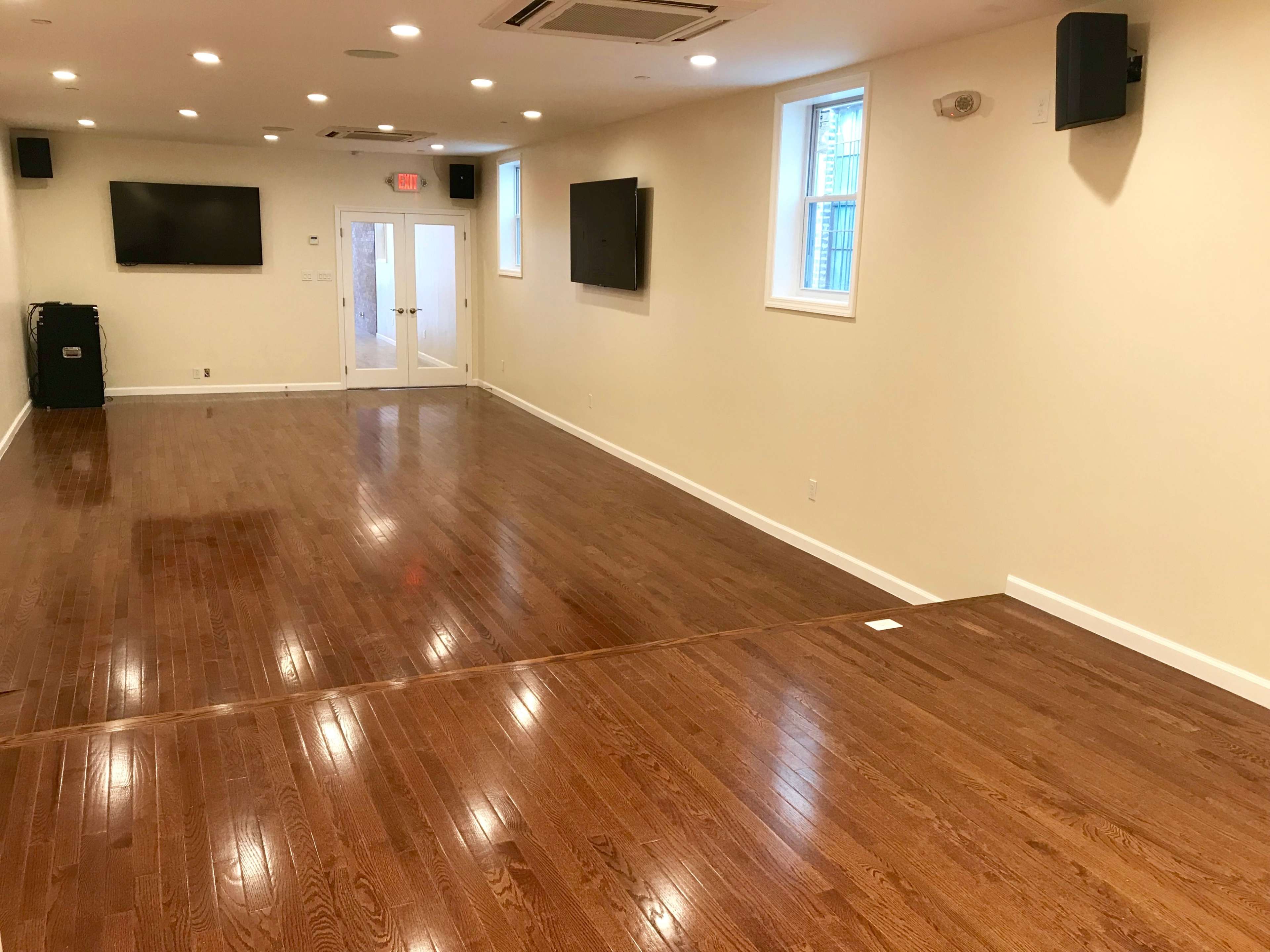 A spacious, empty room with hardwood flooring, two wall-mounted televisions, and large windows.