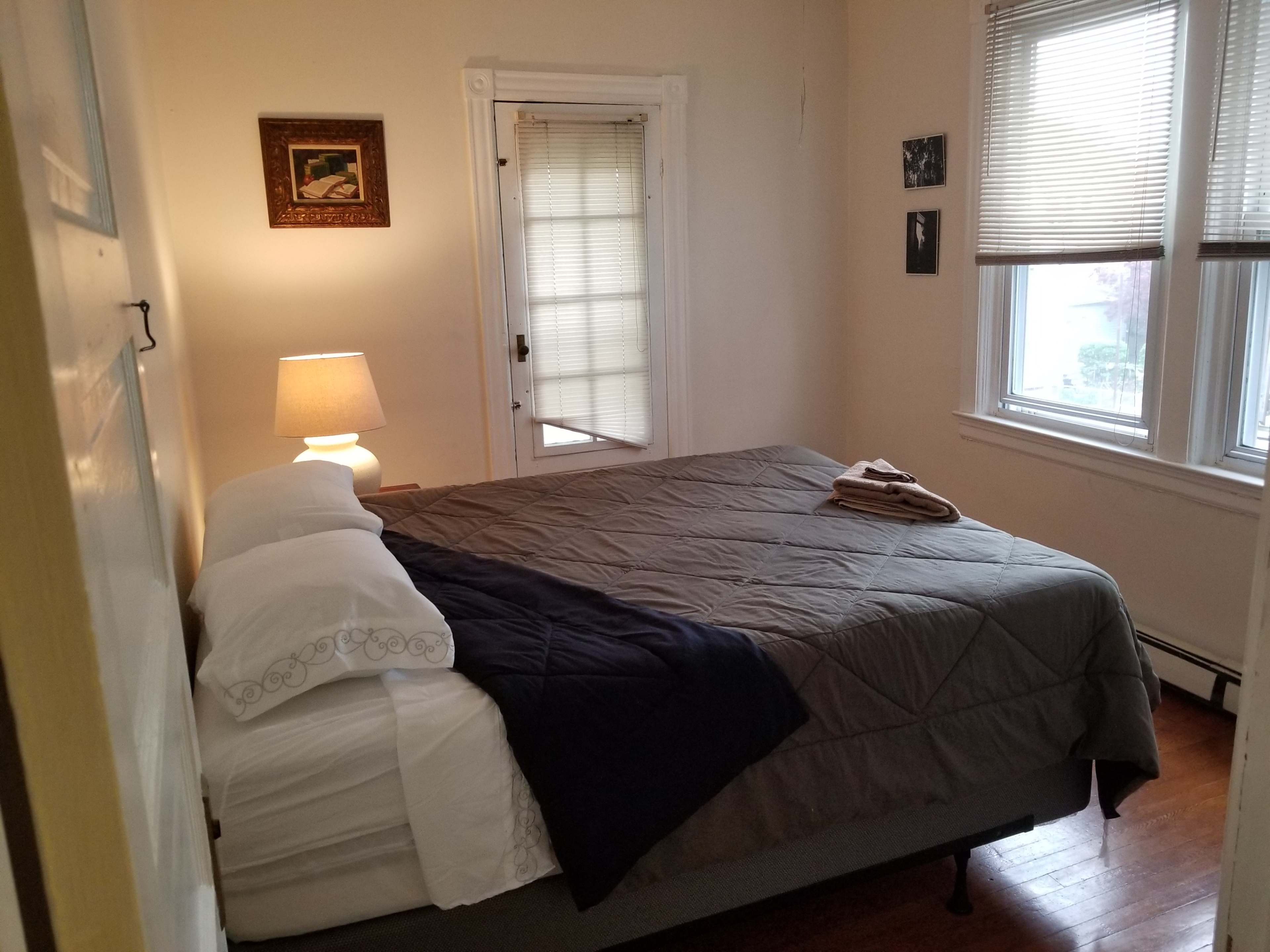 A furnished bedroom with a large bed, a lamp on a bedside table, and a window allowing natural light to enter the room.